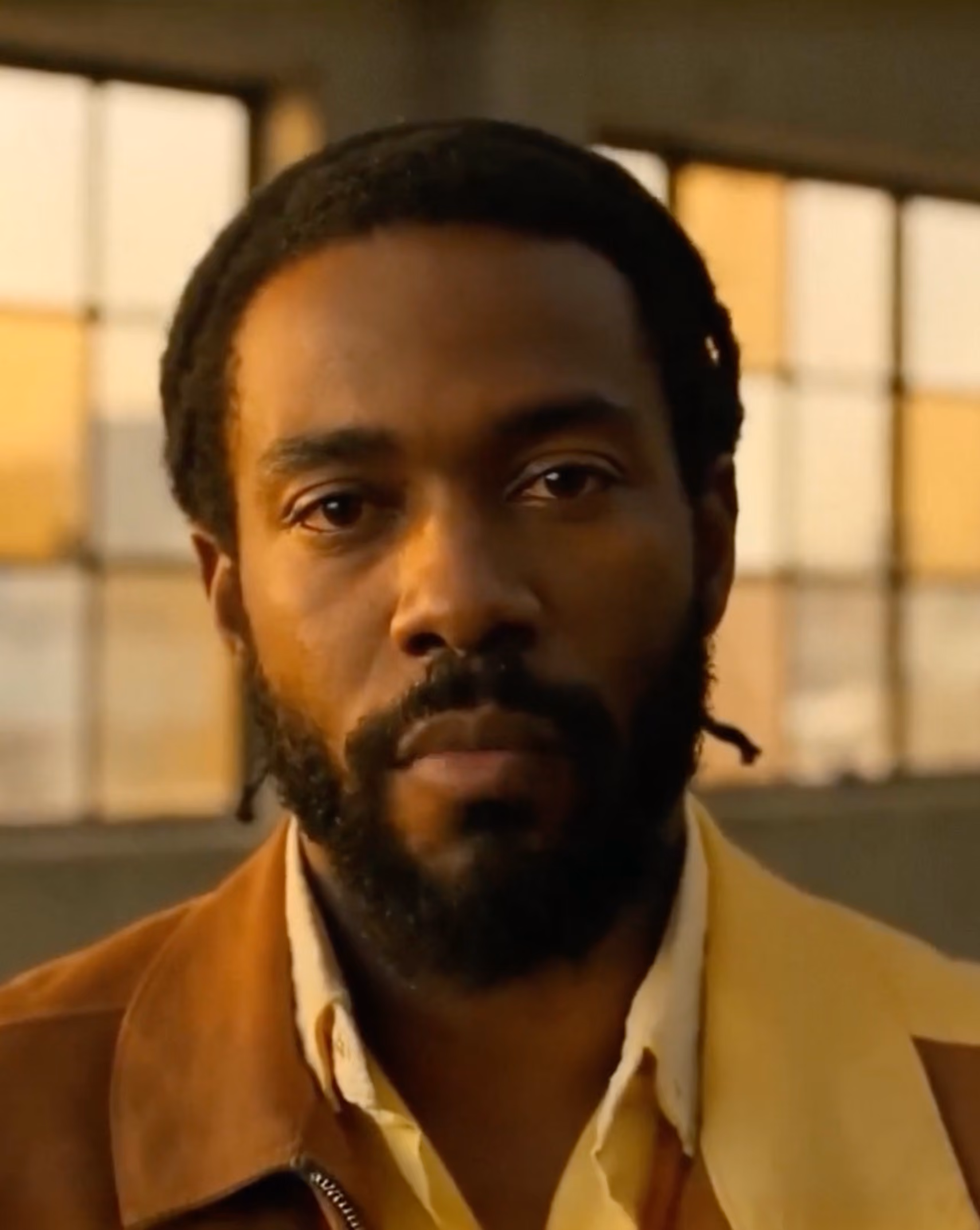 Close-up portrait of a serious man with a beard and braided hair, wearing a brown jacket over a yellow shirt, with a blurred window background.