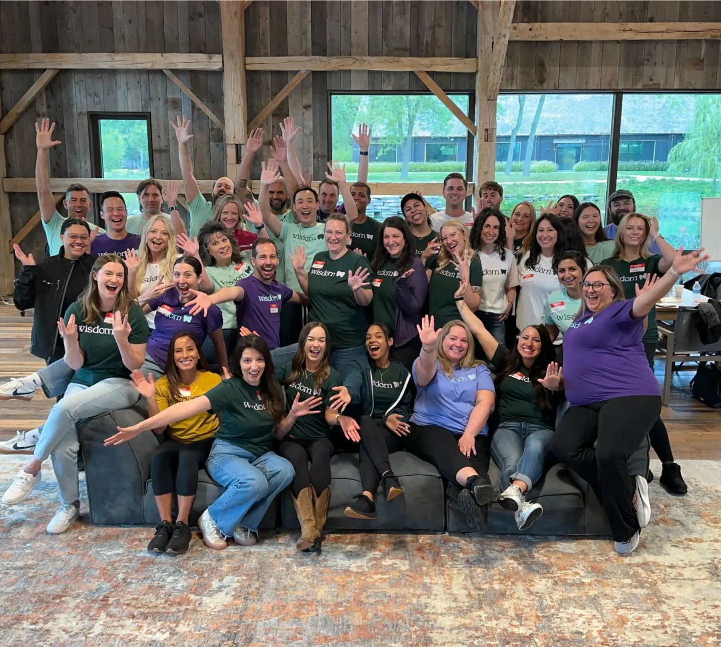 Group of smiling people wearing colorful 'Wisdom' t-shirts posing enthusiastically with raised hands in a cozy room with wooden beams and large windows.
