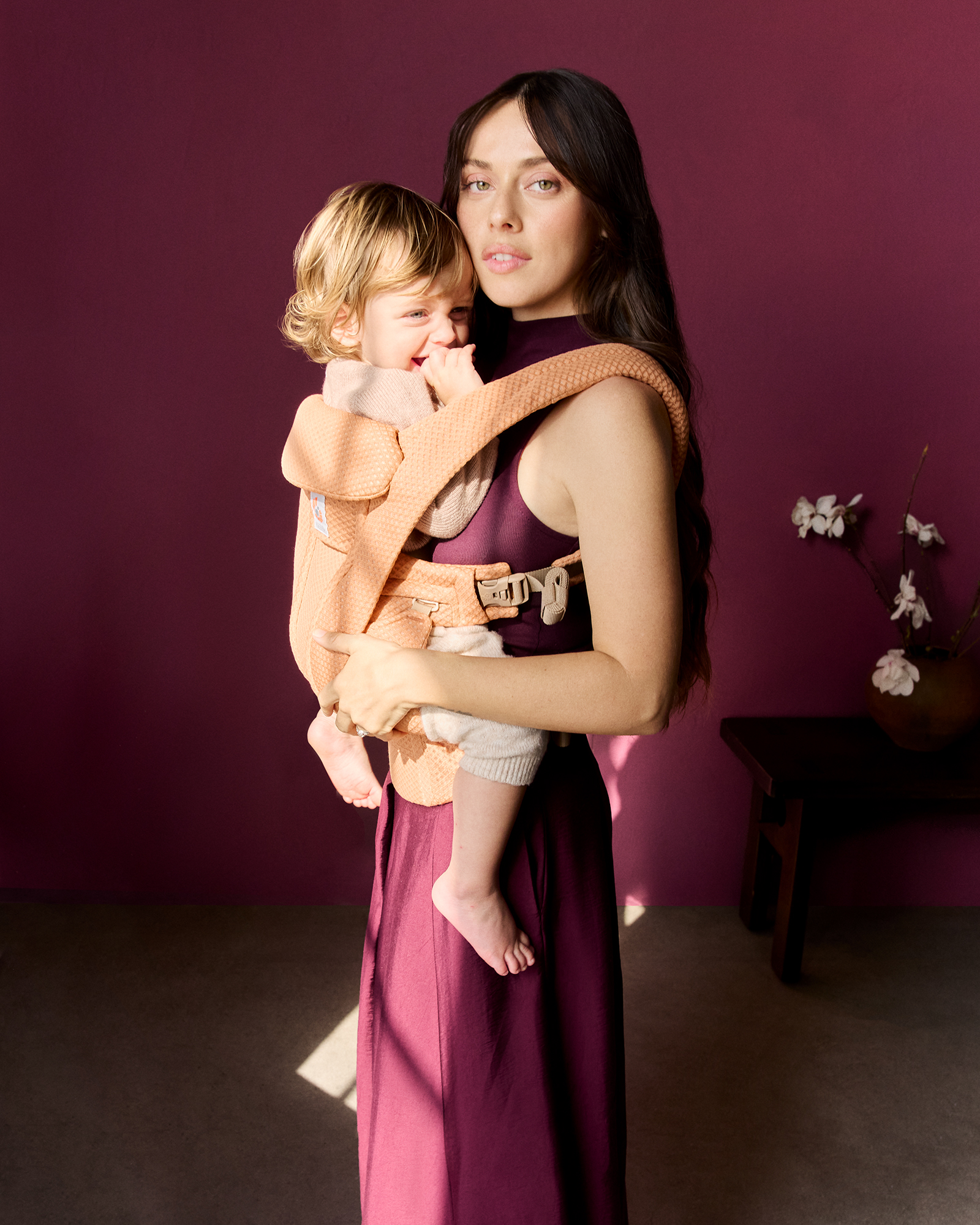 Parent carrying a smiling toddler in a peach Ergobaby mesh carrier