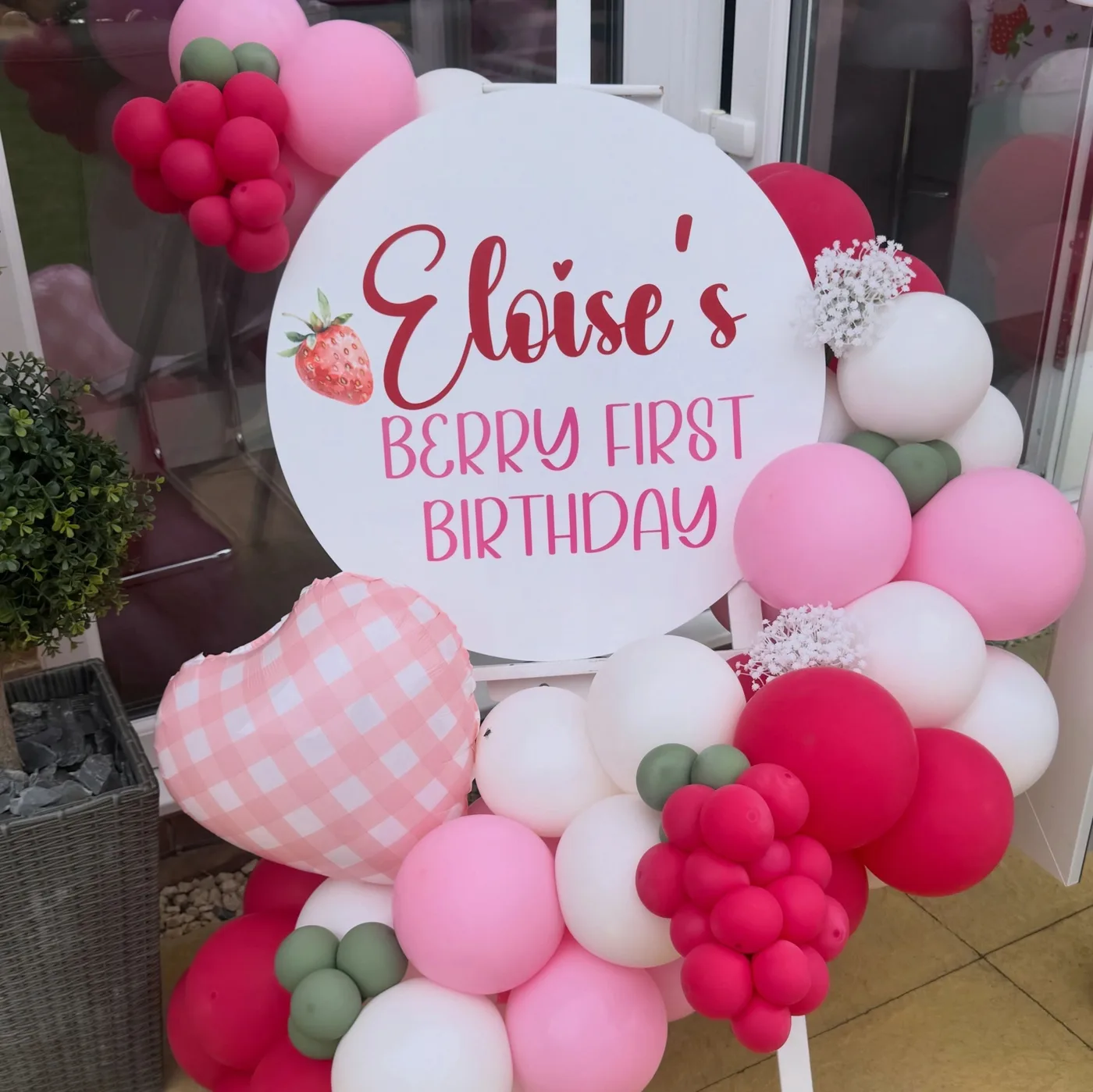 A pink and red strawberry-themed balloon display with a personalised first birthday sign