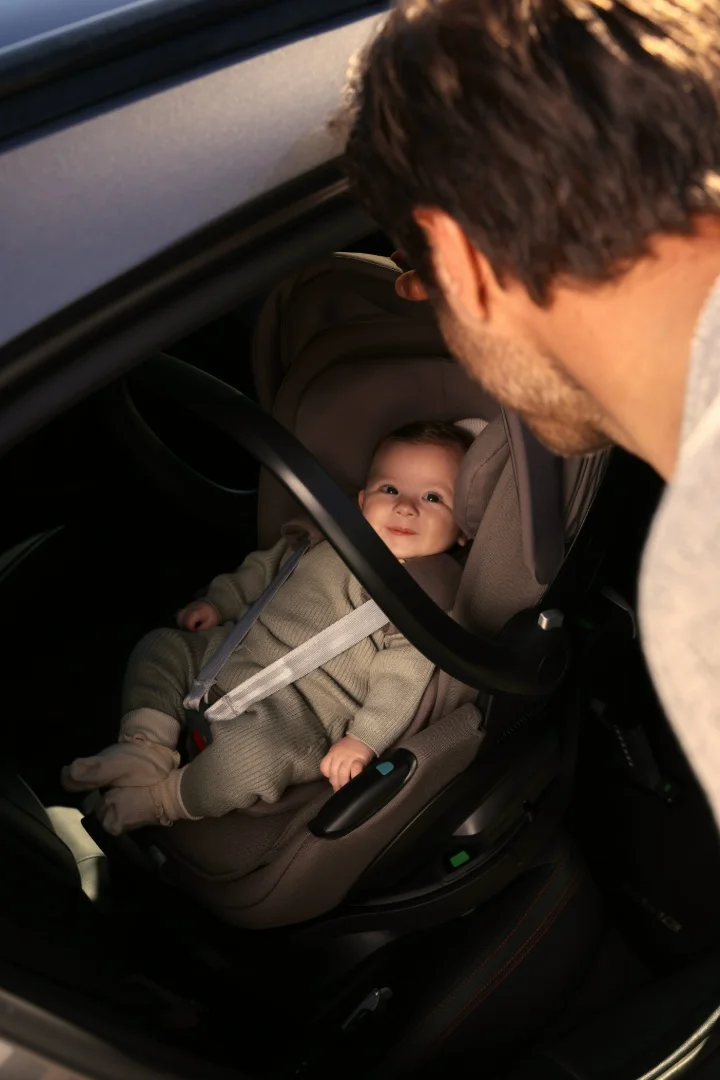 A smiling baby strapped into an Inglesina Darwin infant car seat as a parent leans in