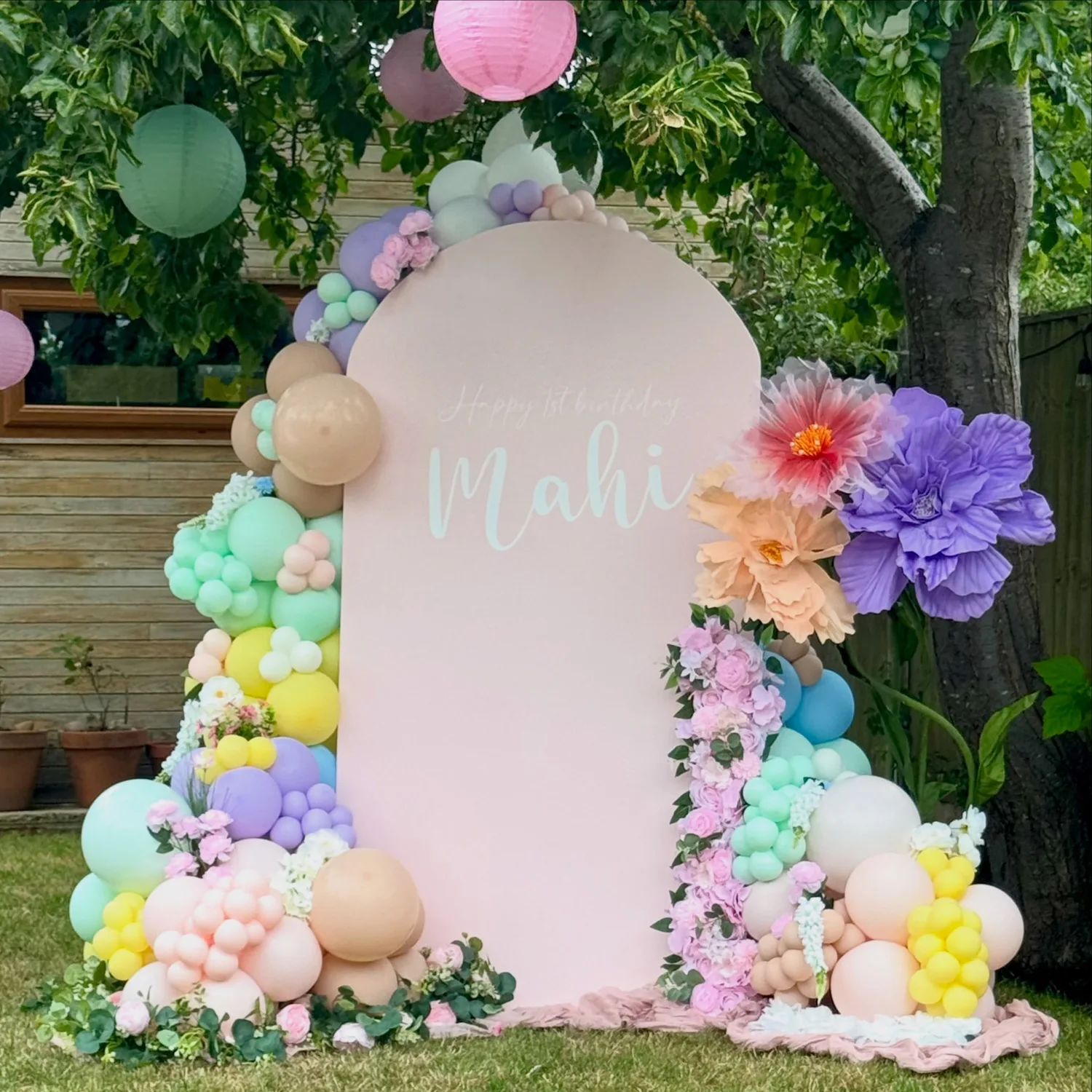 A pastel balloon garland with paper flowers around a personalised arched sailboard backdrop by Posh Tots Play