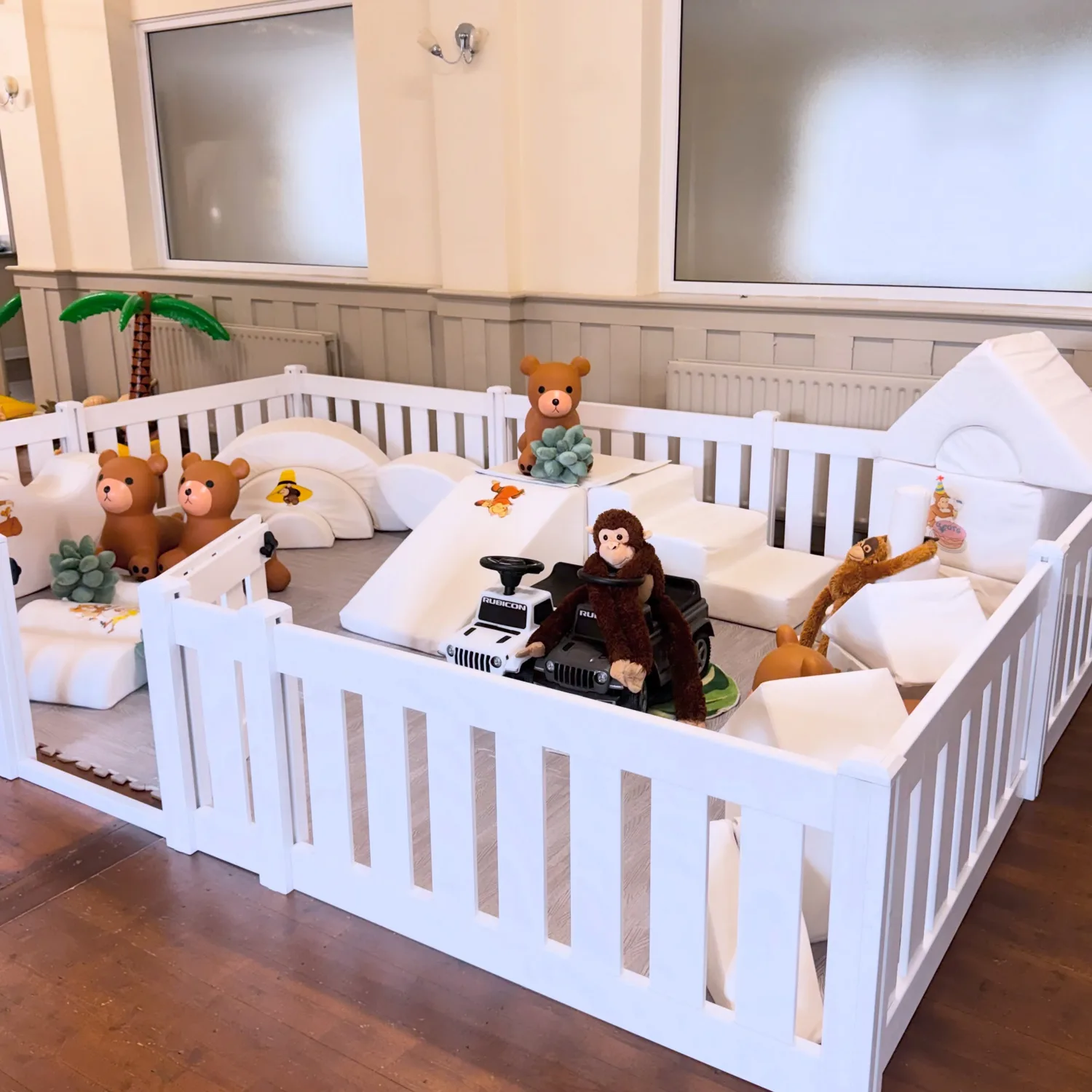 A white fenced soft play area with shapes, a slide, ride-on cars and teddies by Posh Tots Play