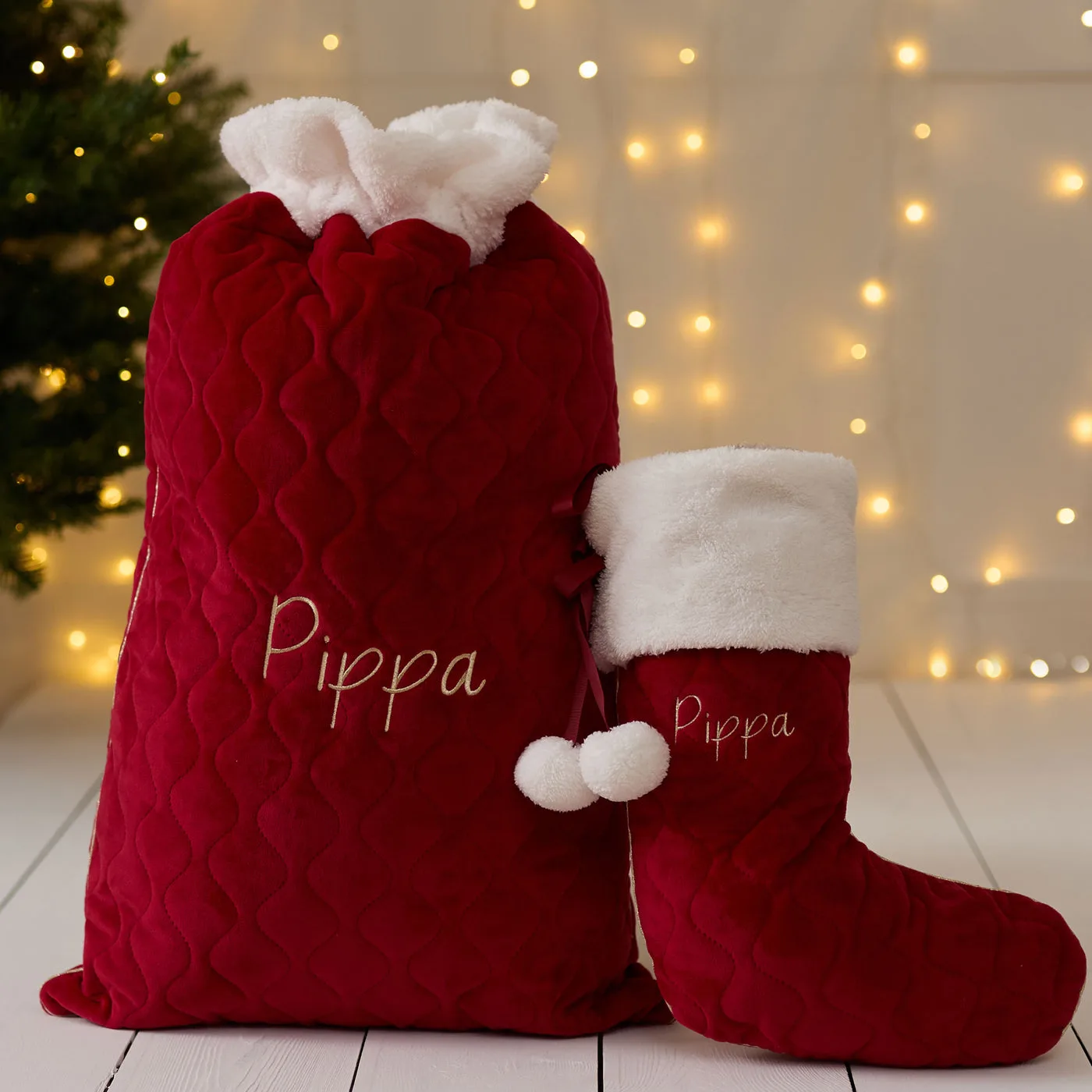 A red quilted velvet Christmas sack and matching stocking with white faux fur tops, each embroidered with the name Pippa in gold, beside a Christmas tree with fairy lights