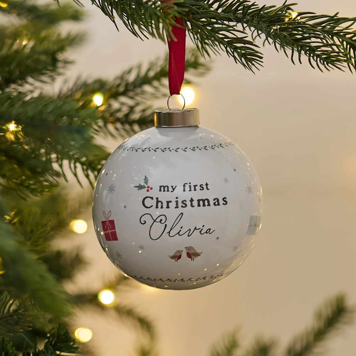A white ceramic My First Christmas bauble personalised with the name Olivia, hanging from a red ribbon on a Christmas tree