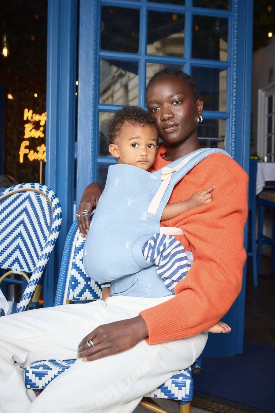 A mum sitting at a cafe with her baby in a pale blue Tula Original carrier in the Covey cotton print