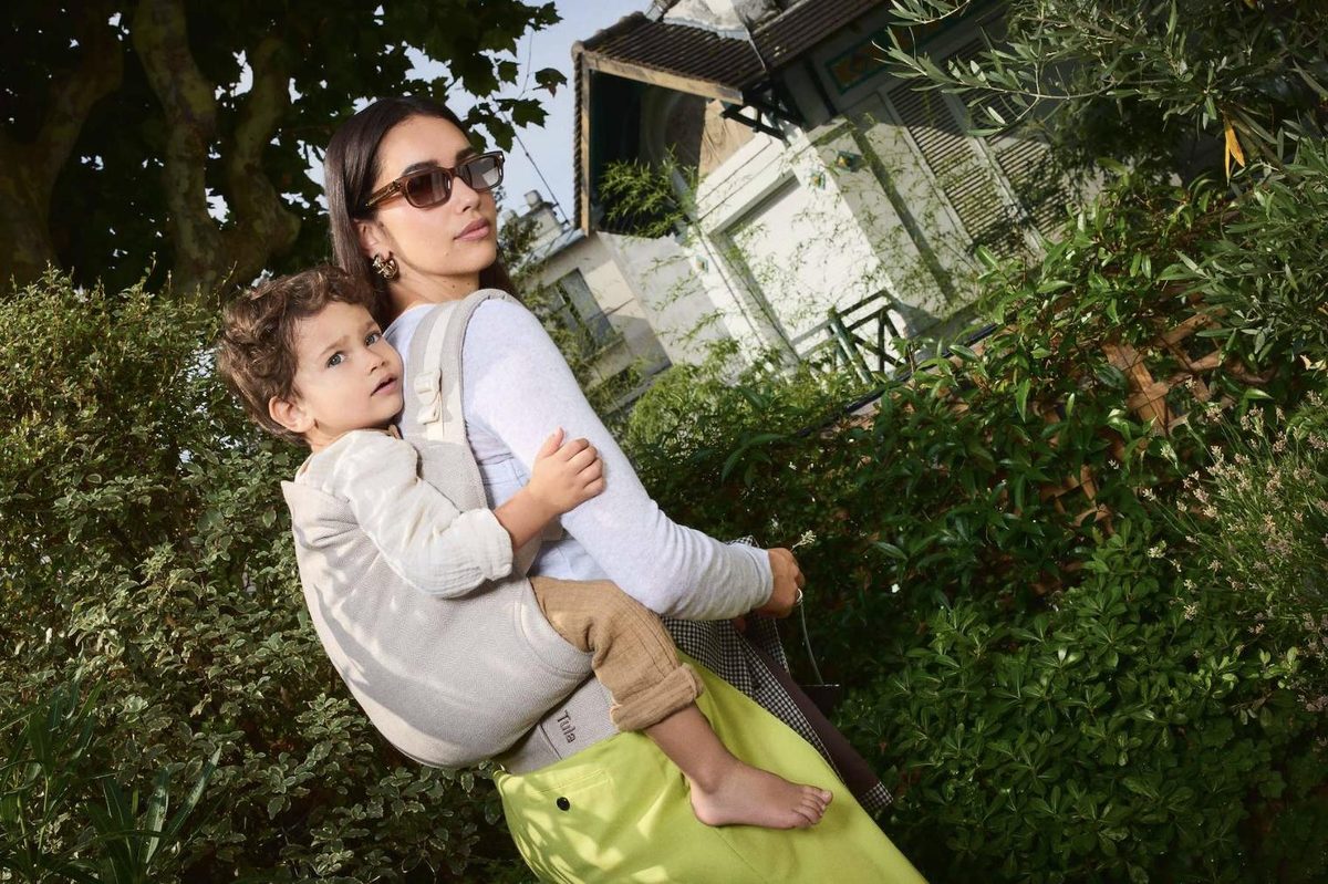 A mum carrying her toddler on her front in a Tula Original carrier in Sand herringbone linen in a garden