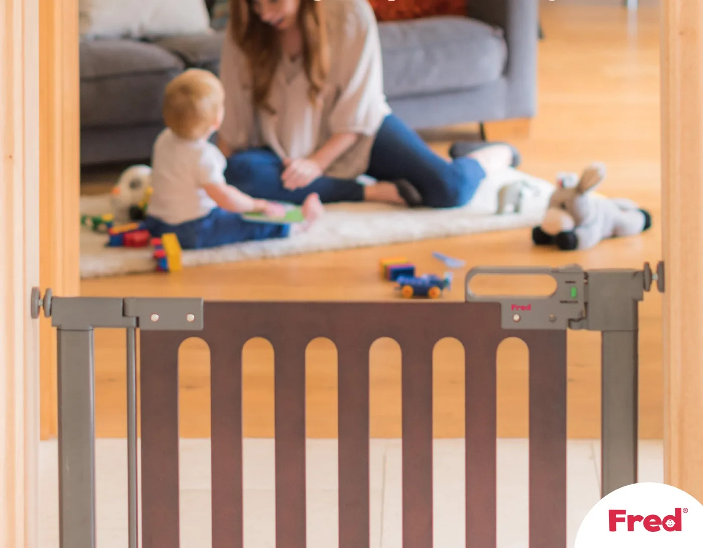 A mum sits on a living room rug playing with her baby among scattered toys, seen through a grey and wood safety gate fitted across the doorway