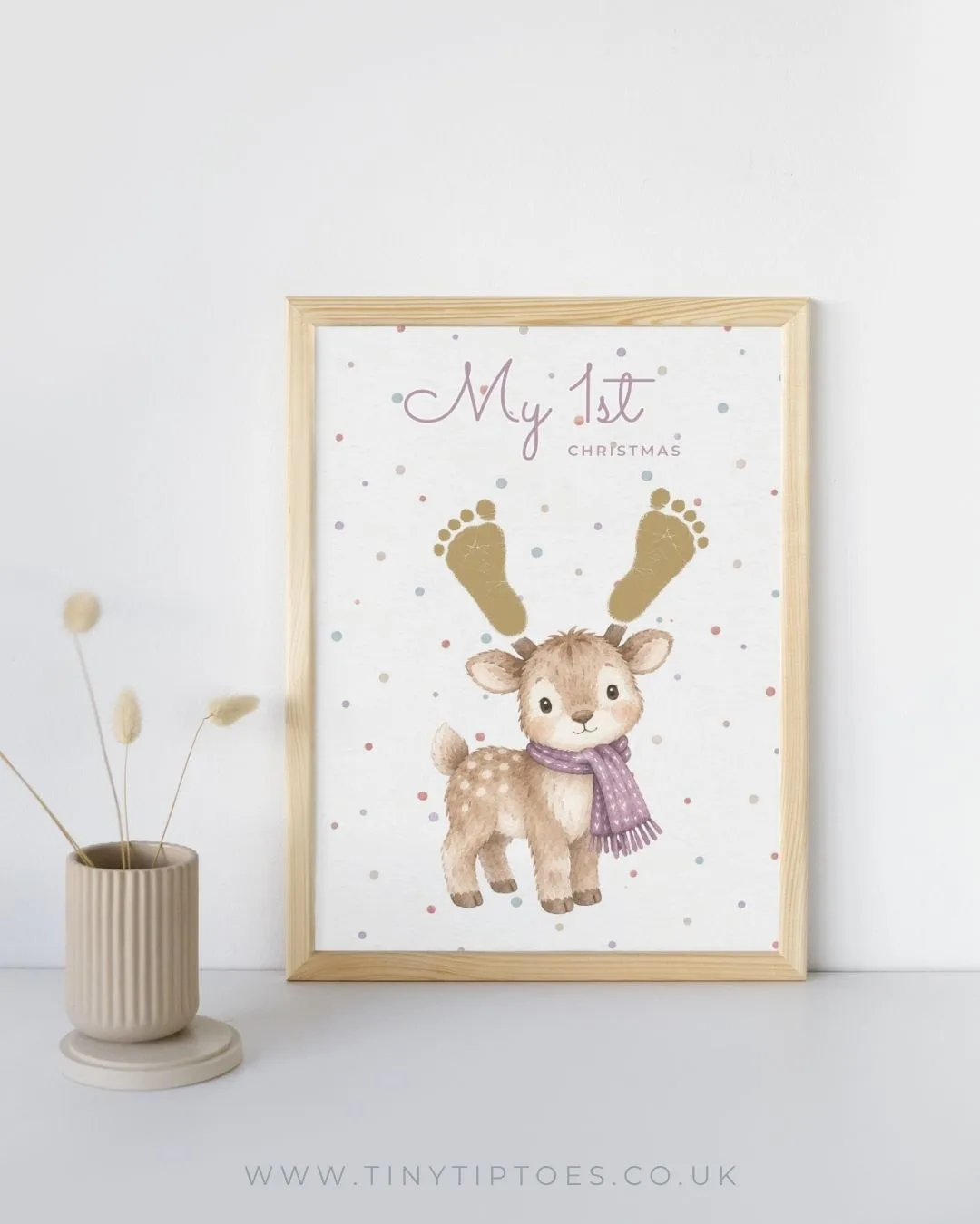 A framed 'My 1st Christmas' reindeer print where the antlers are made from two gold baby footprints, propped against a white wall beside a vase of dried flowers