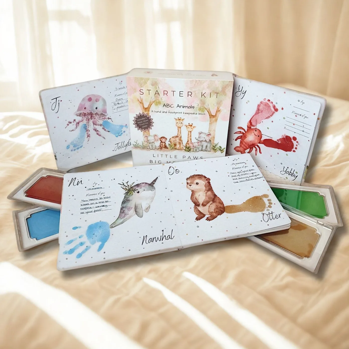 The ABC Animals keepsake book open on a bed with hand and footprint animal illustrations, including a crab and a narwhal, next to the starter kit box and coloured ink pads