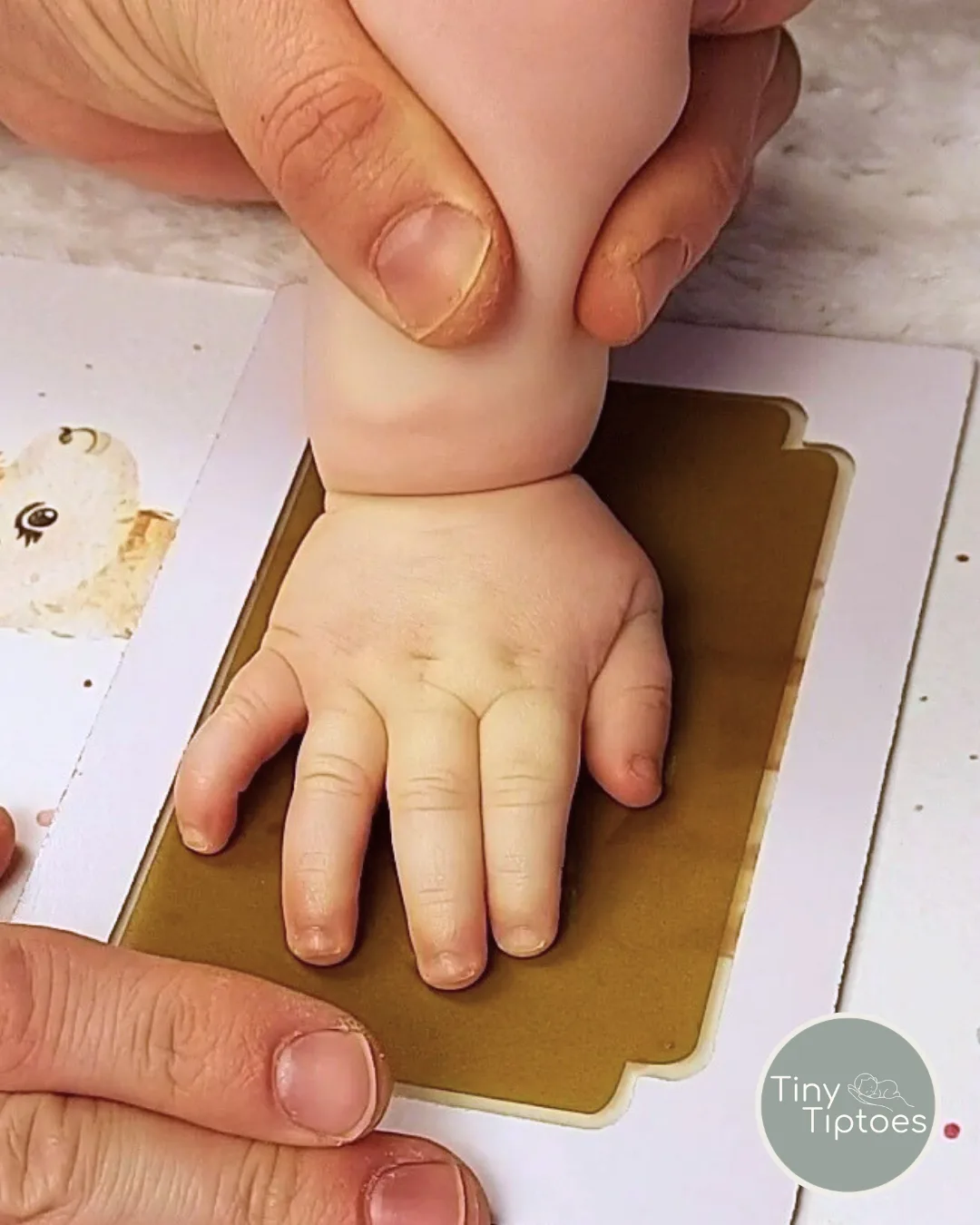 A grown-up's hands gently pressing a baby's hand onto a gold clean-touch ink pad laid over a printed keepsake