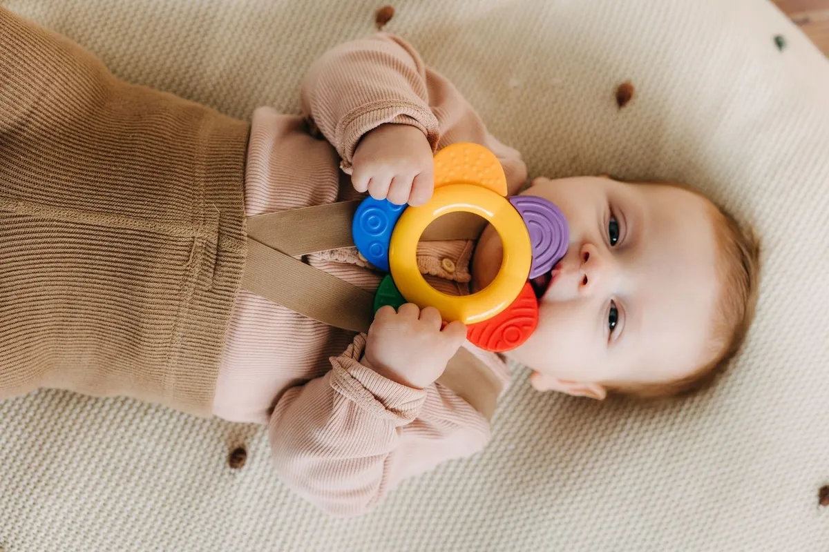A baby lying down chewing on a colourful teething toy