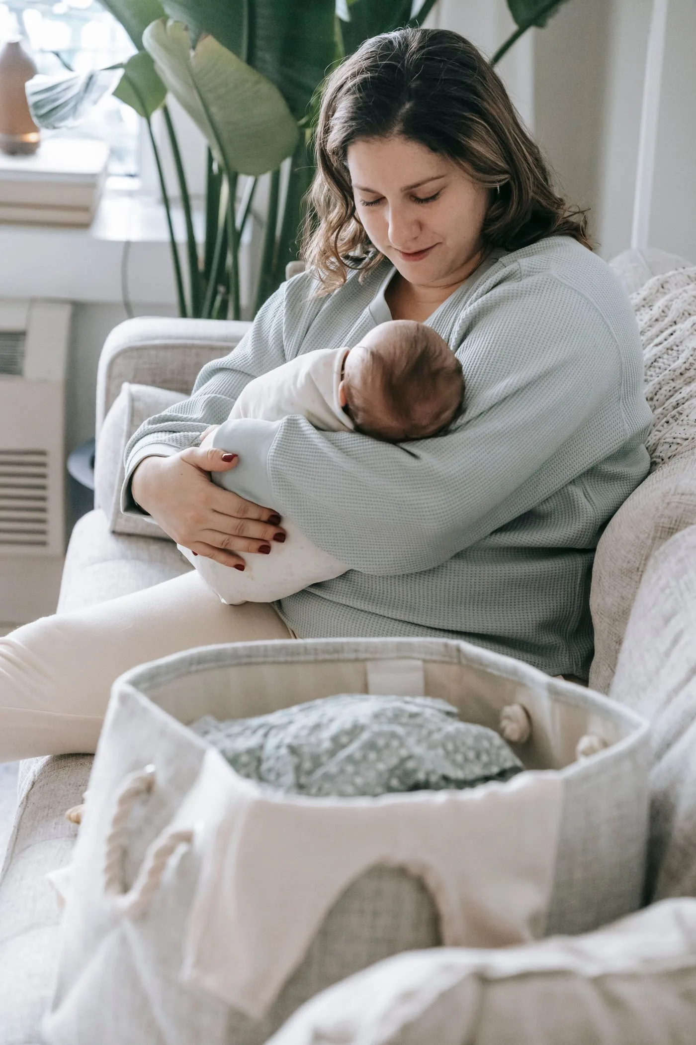A new mum sitting on the sofa at home holding her newborn, with a basket of laundry beside her