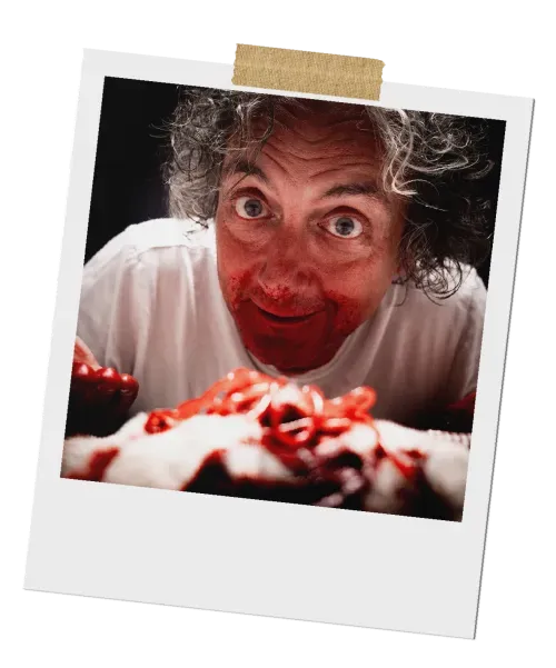 A Polaroid photo of a man with wild, graying hair, looking shocked with wide eyes. A large knife is held in his mouth, dripping with fake blood.