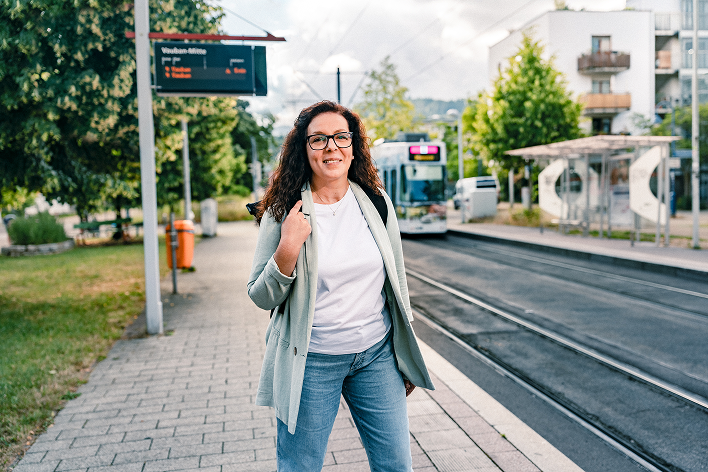 Eine lächelnde Haufe Group Mitarbeiterin mit Rucksack steht an einer Straßenbahnhaltestelle.