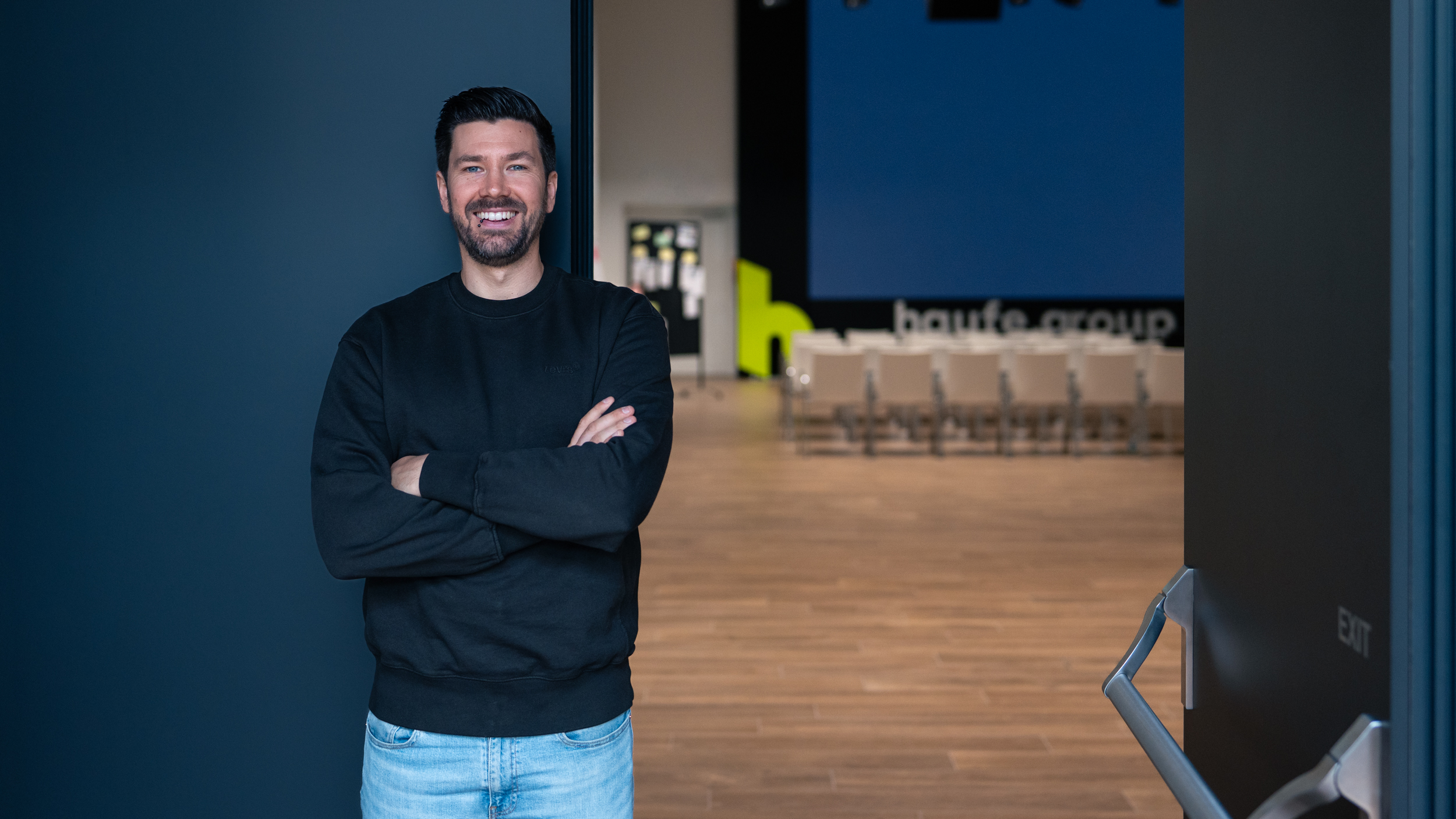 Mathias Wohllaib steht mit verschränkten Armen vor den Eingangstüren des Veranstaltungsraum "The Cube" der Haufe Group. Er lächelt in die Kamera.