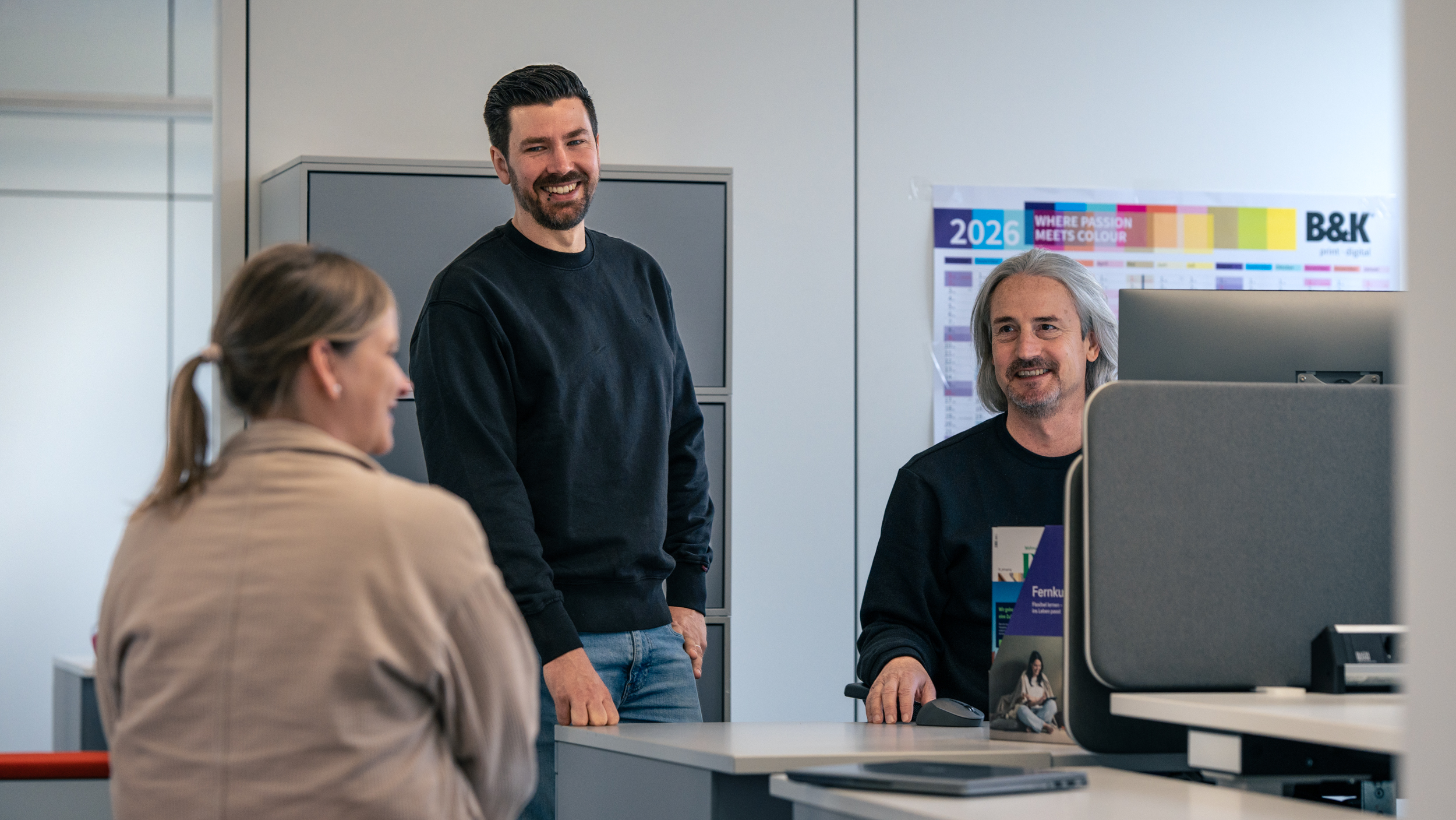 Mathias steht hinter einem Schreibtisch in einem hellen Büro. Ein Kollege und eine Kollegin sitzen links und rechts neben ihm. Die drei sind in einem Gespräch.