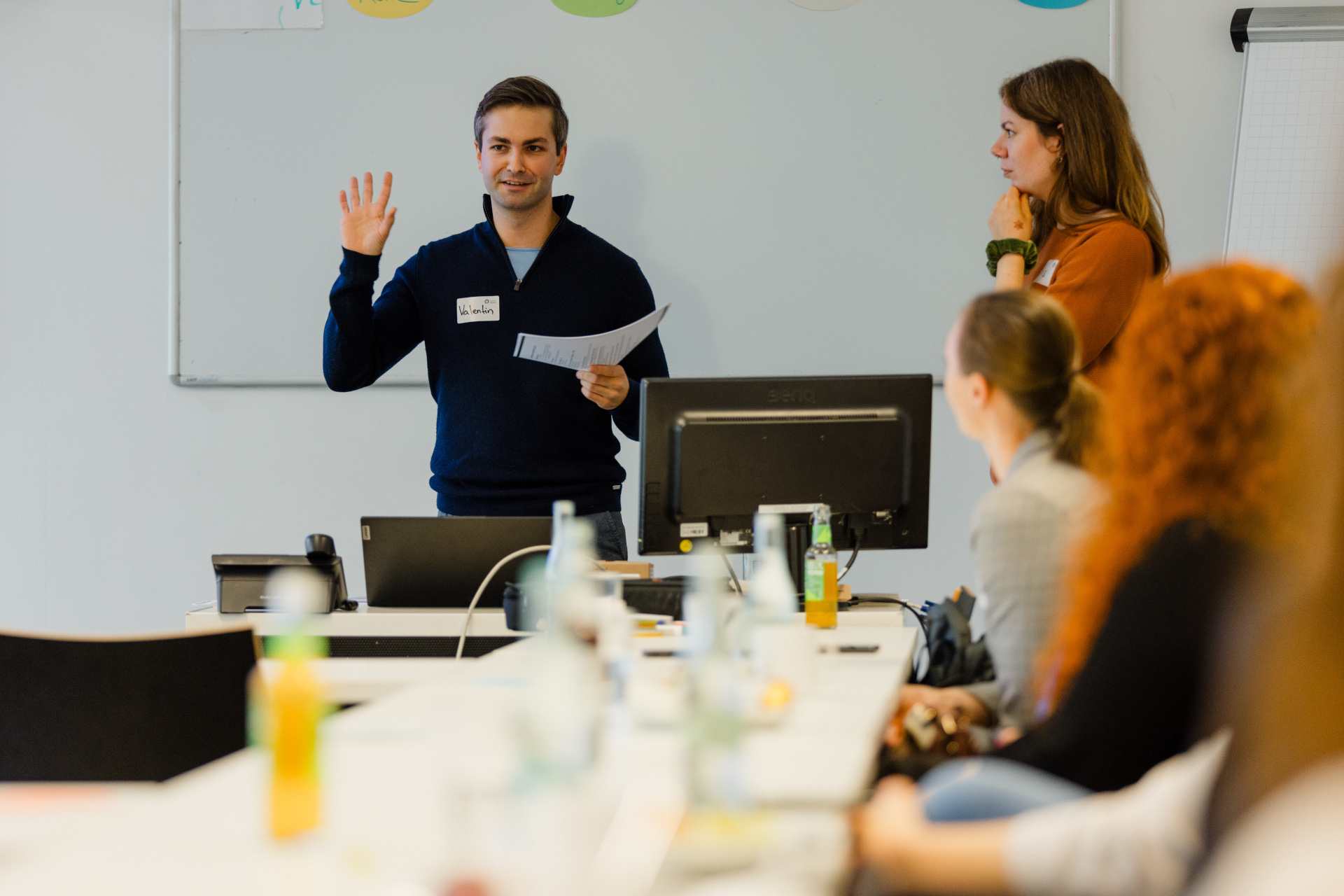Valentin Burger steht in einem Workshop-Raum und spricht zu den Teilnehmenden. In der Hand hält er einen Zettel mit Notizen. Rechts im Bild sind Teilnehmende zu sehen, die ihm zugewandt sitzen und zuhören.