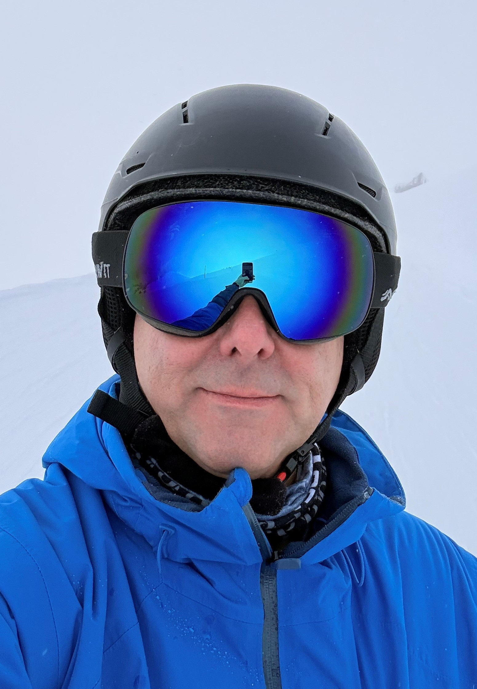 Torsten trägt einen Skihelm sowie eine blaue Skibrille und einen blauen Skianzug und fotografiert sich vor einer vernebelten verschneiten Piste.