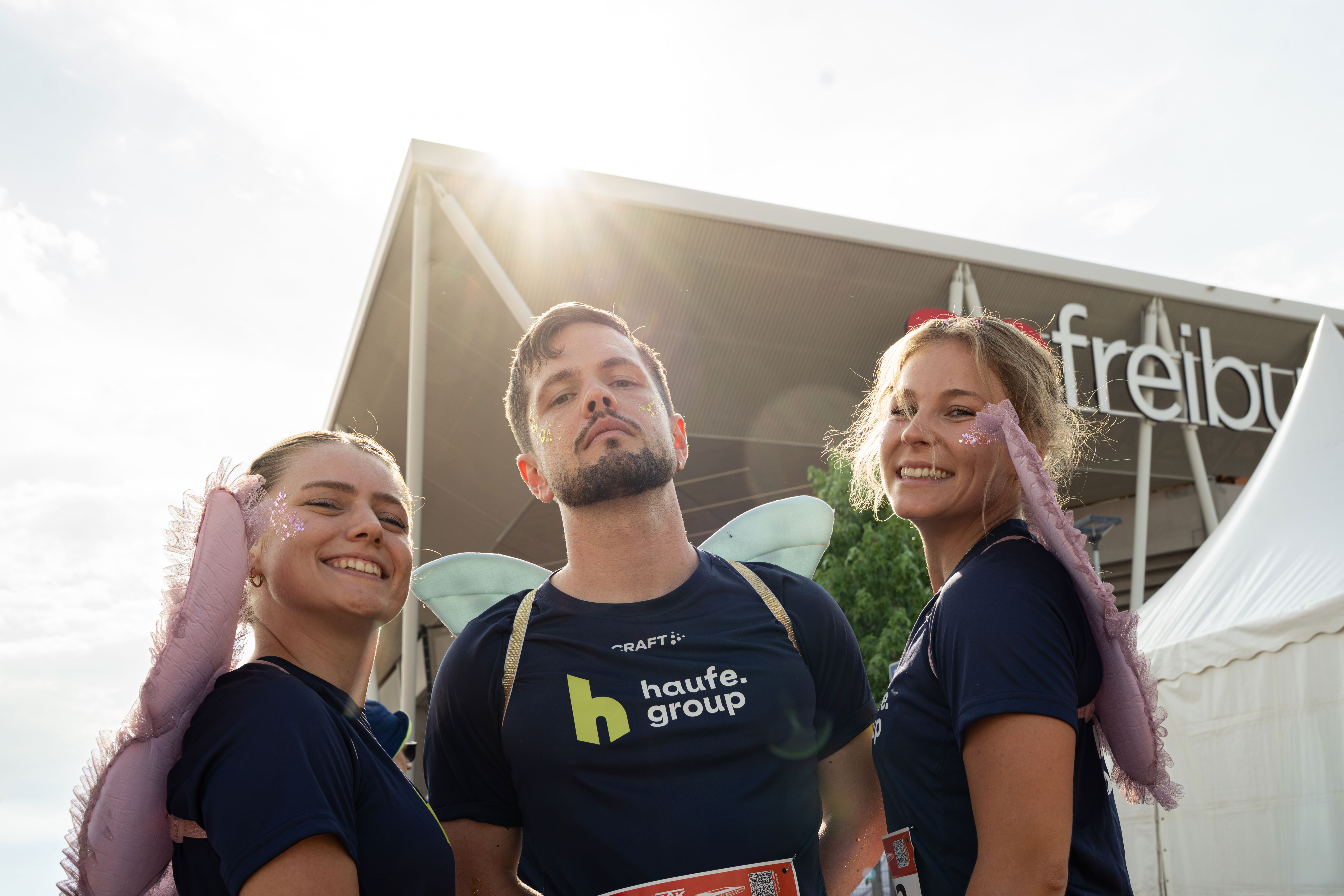 Cindy Koller, Gregory Rech und Luisa Licht stehen vor dem SC-Stadion in Freiburg. Sie tragen Haufe Group-Trikots, sind mit Glitzer geschminkt und haben Flügel auf dem Rücken.