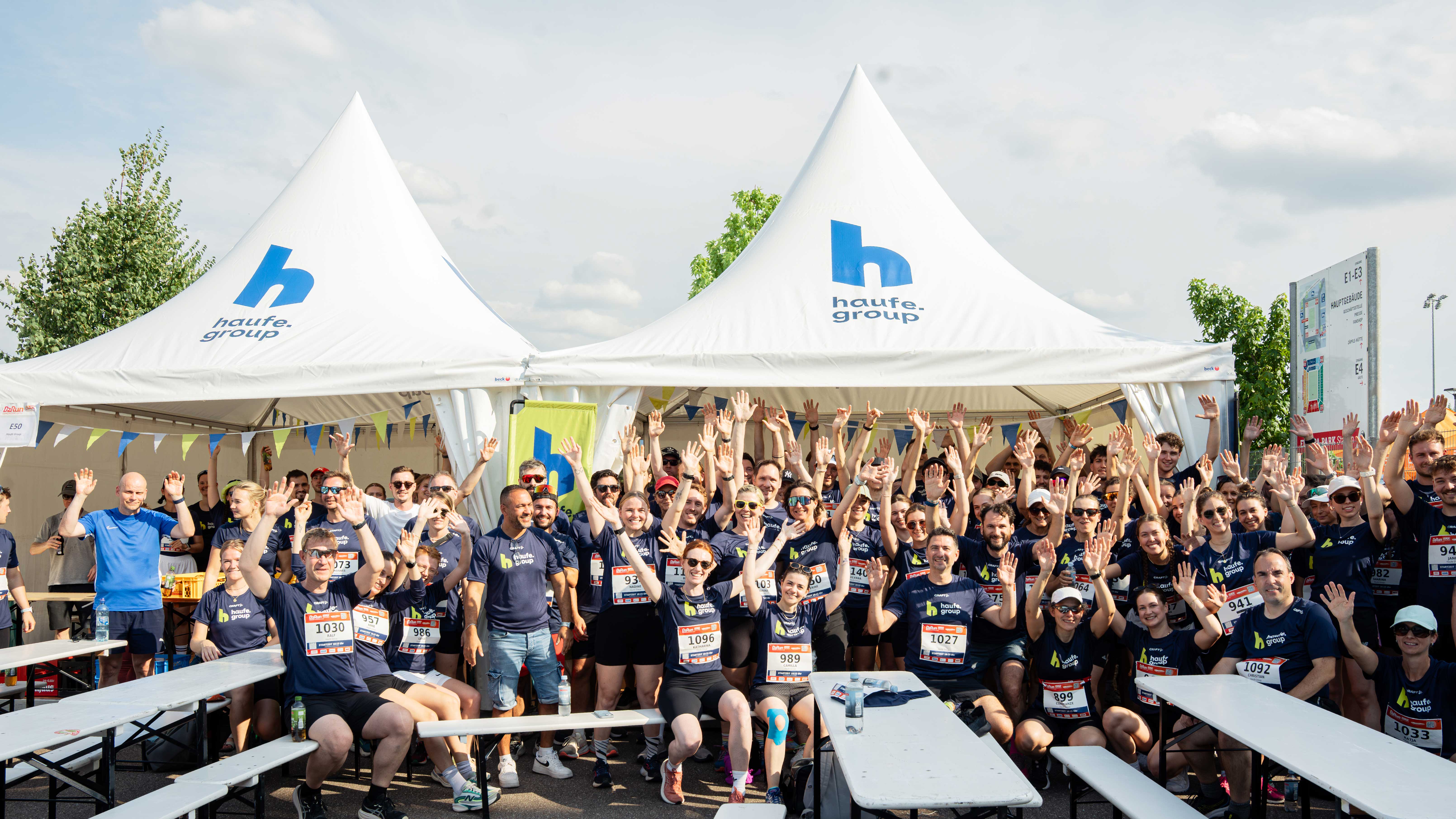 Zahlreiche Mitarbeitende in Haufe Group-Trikots stehen vor dem Zelt der Haufe Group beim B2Run in Freiburg und reißen die Hände in die Höhe.