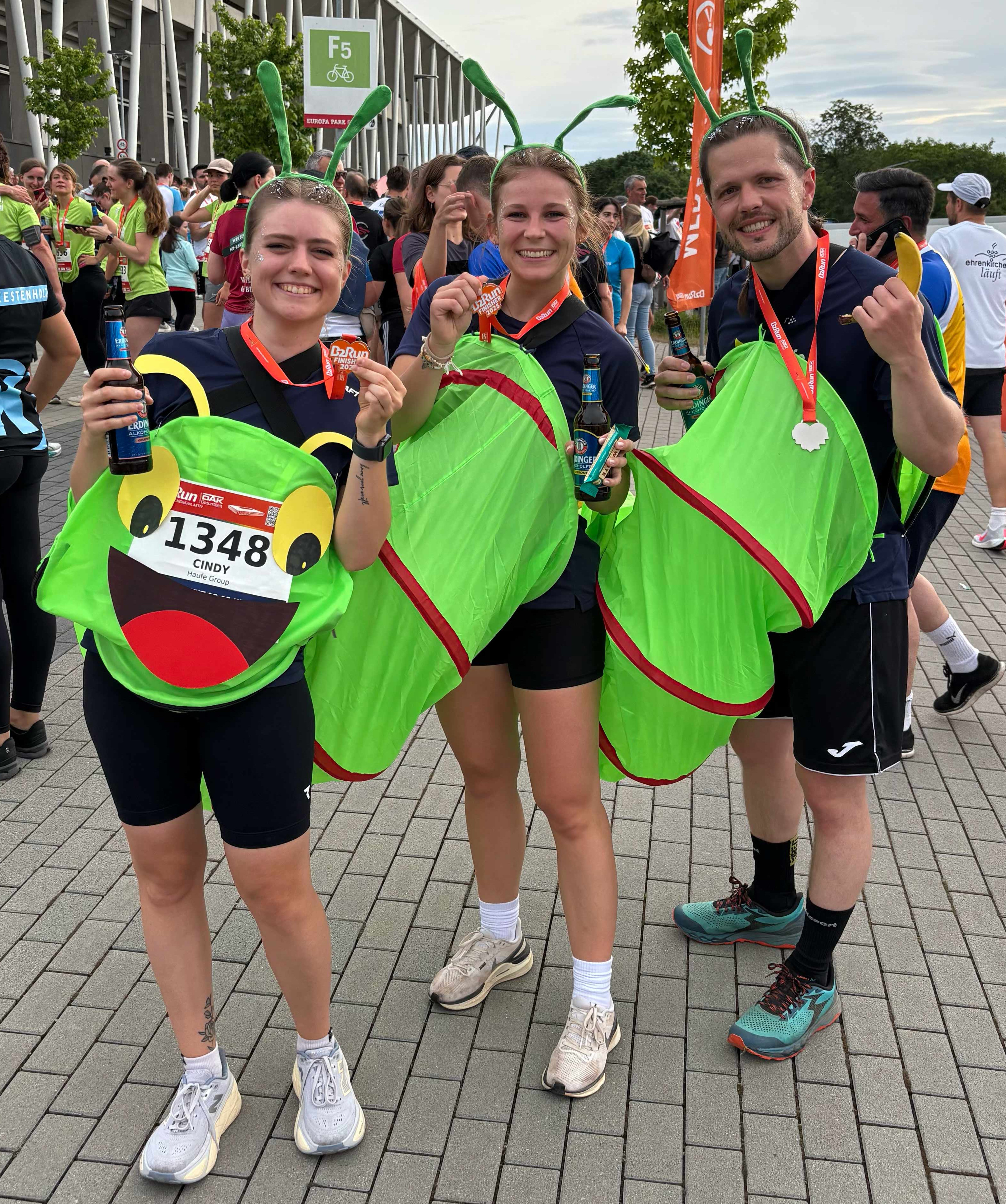 Cindy, Luisa und Gregory stehen im Raupenkostüm und in Haufe Group-Trikot vor der Messe Freiburg und halten ihre B2Run-Medaille in die Kamera.