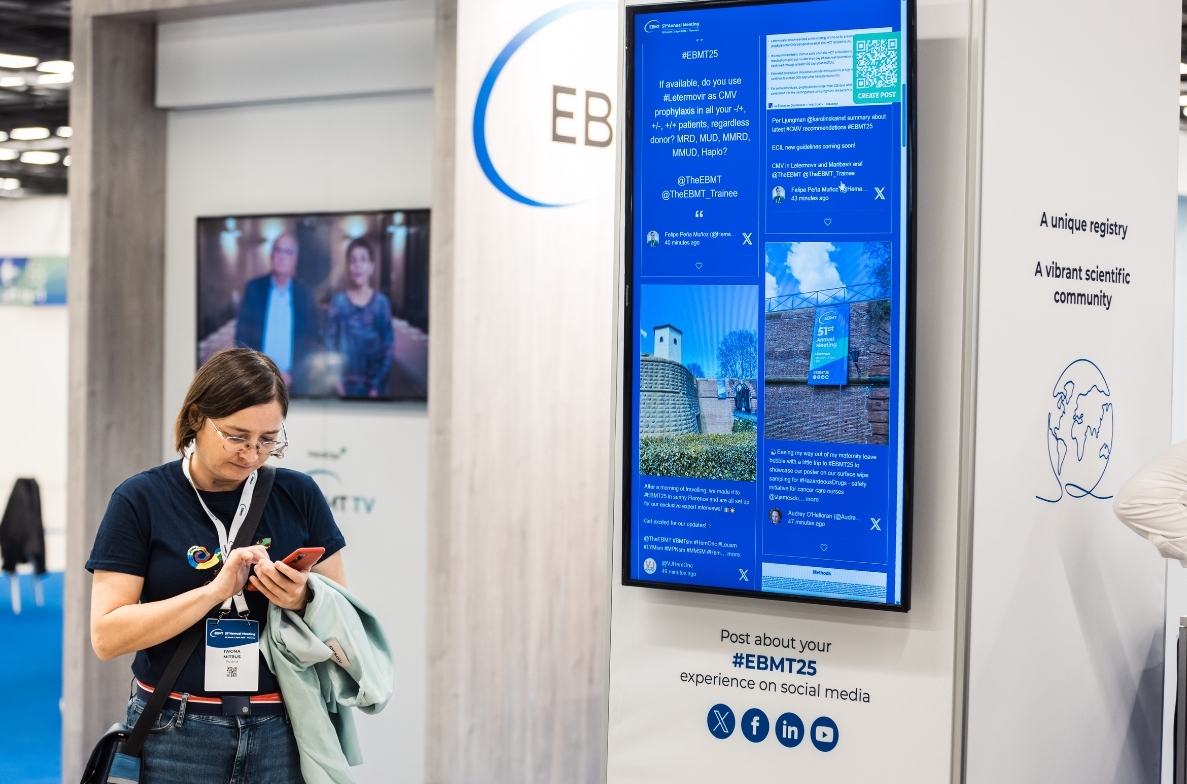 Eine Frau steht auf einem Fachkongress vor einer digitalen Social Wall und schaut auf ihr Smartphone. Auf dem großen Bildschirm werden Social Media Beiträge mit dem Hashtag #EBMT25 angezeigt.