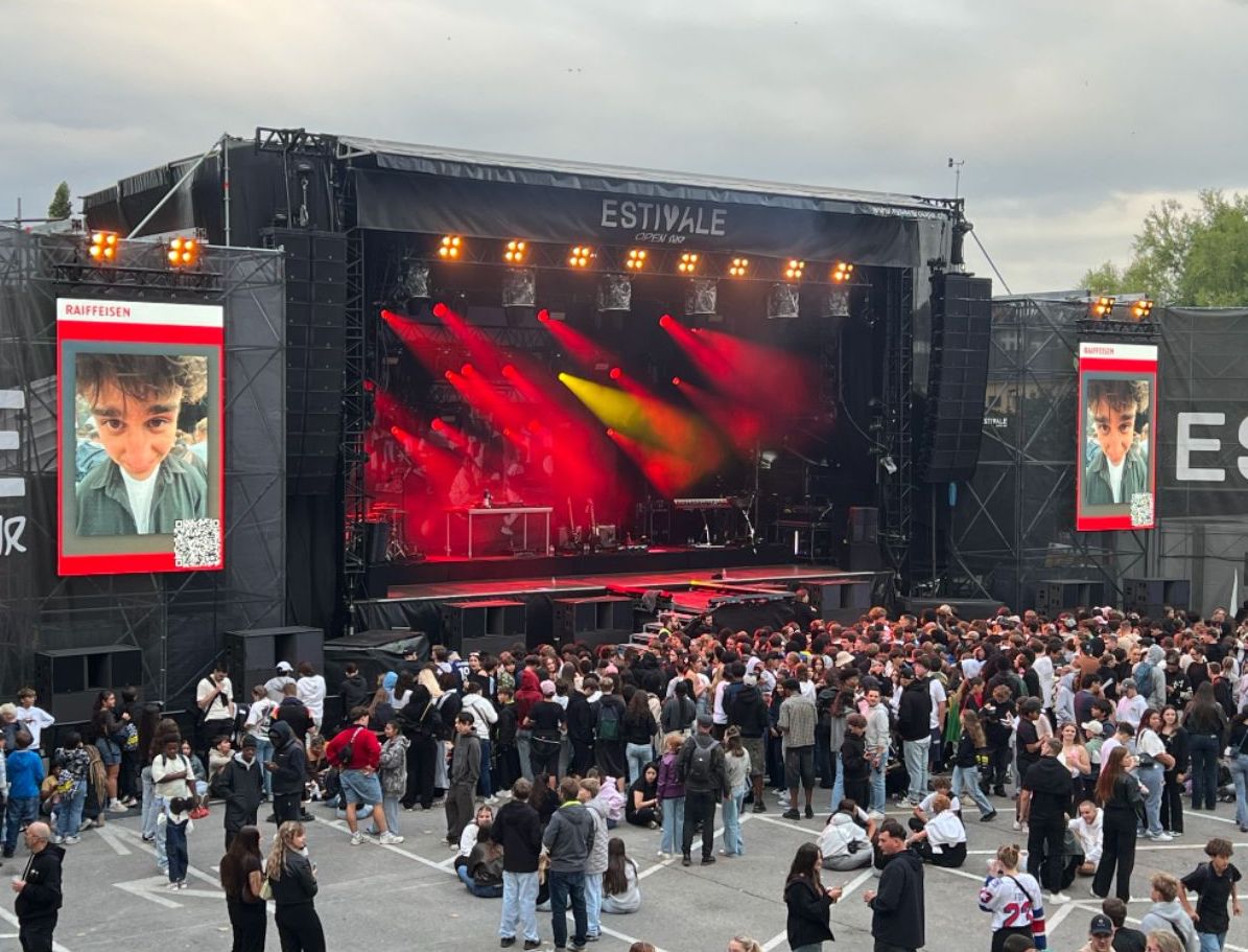 An open air music festival. Attendees are mostly yourn adults, they are looking at the stage. On the right and left side of the stage is a social wall displaying UGC from attendees and a QR code for anyone to scan and post photos.