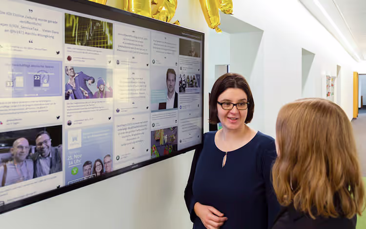Zwei Personen stehen vor einem Fernseher, auf dem eine LinkedIn-Social-Wall angezeigt wird.