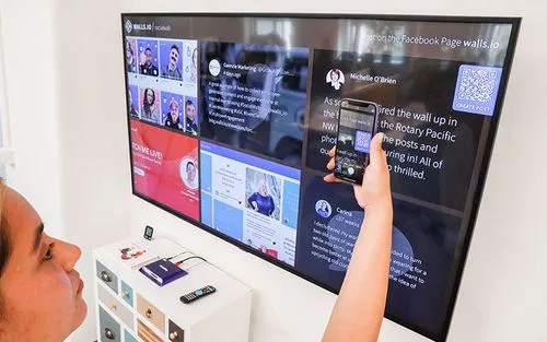 Eine Frau scannt einen QR-Code auf einer digitalen Anzeige mit einer Social-Media-Wall. Sie scannt den Code, um Inhalte auf die Wall hochladen zu können.