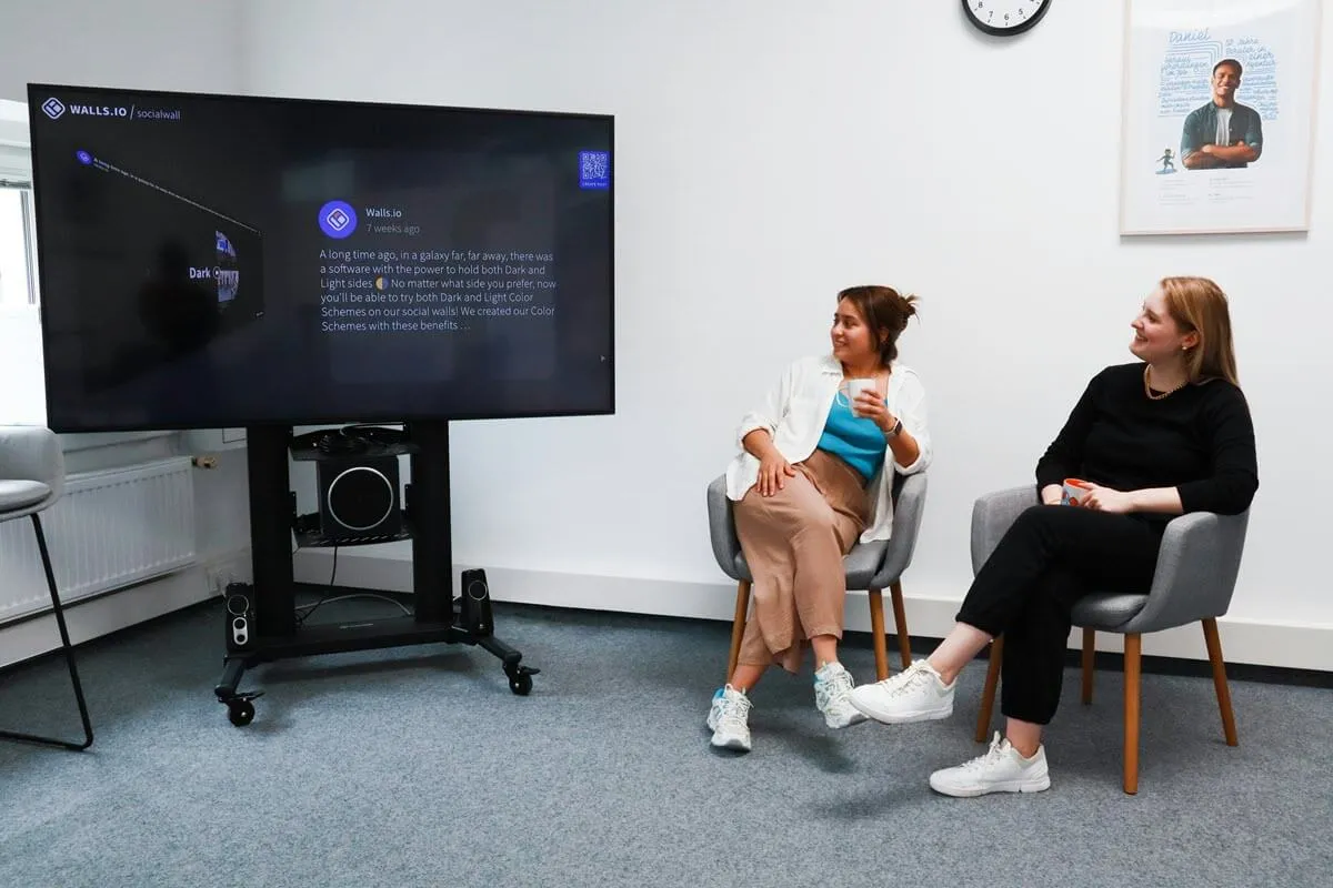 Digital-Signage-Inhalte auf einem Social-Media-Bildschirm in einem Büro; zwei Frauen sitzen auf Stühlen und schauen ihn an.