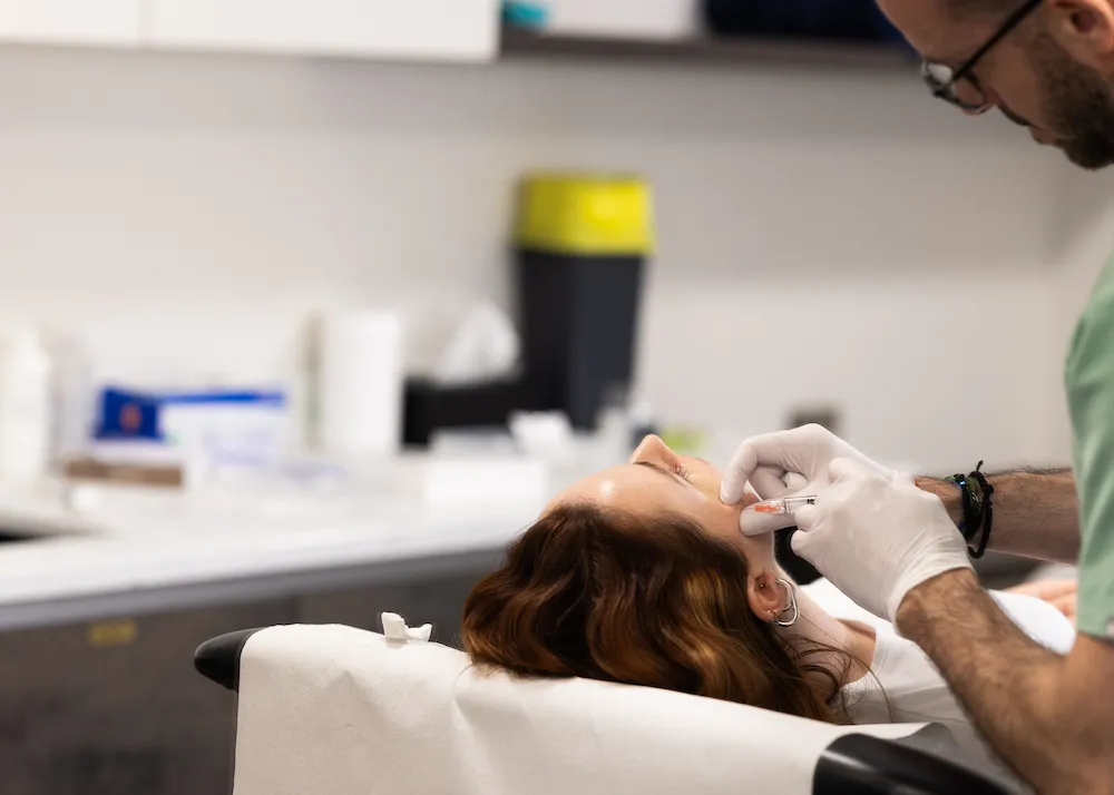Medical professional wearing gloves administering an injection to a woman's cheek in a clinical setting.