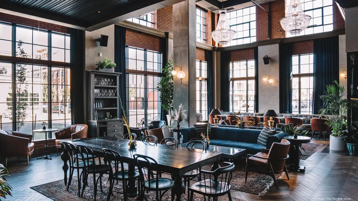 A spacious, elegant hotel lobby with high ceilings, large windows, chandeliers, and a long dining table.