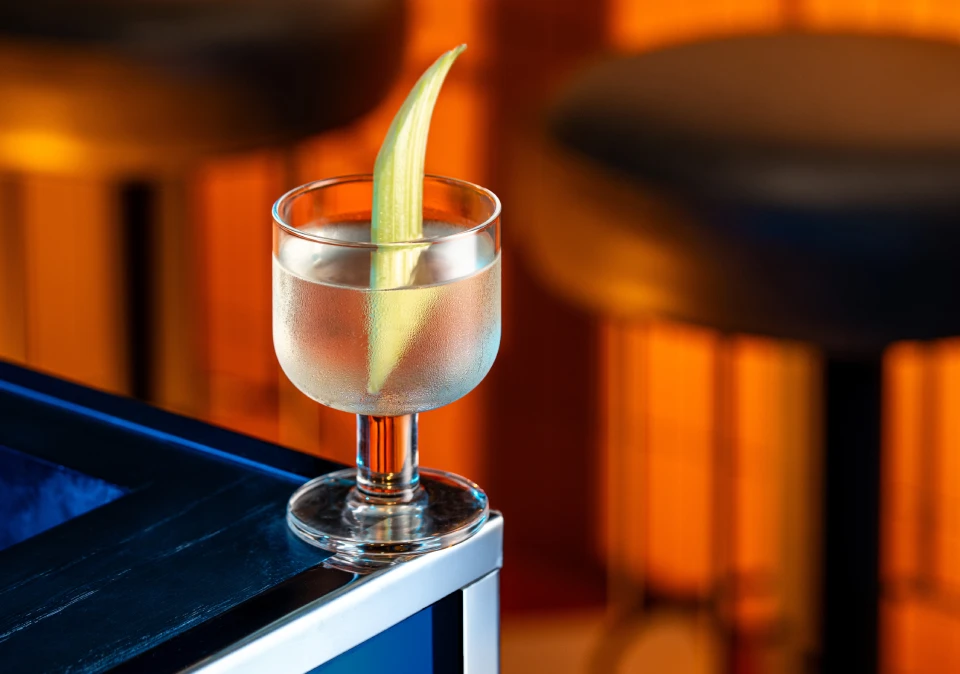A clear cocktail with a celery stick garnish sits in a stemmed glass on a bar counter.