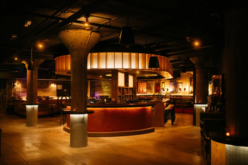 A dark, dimly lit room featuring a circular bar with ambient light and several pillars.