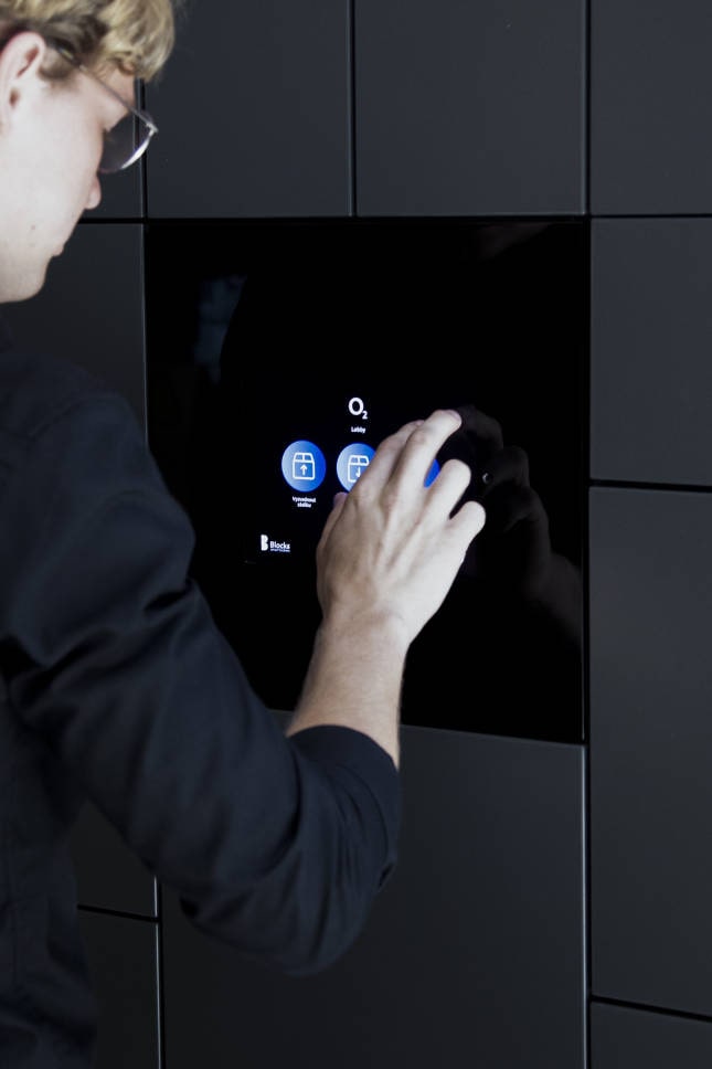 Person interacting with a touchscreen interface embedded in a black smart locker system, selecting options under the label O2 Lobby.