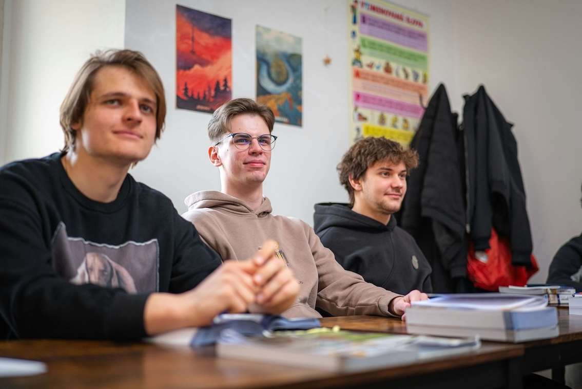 Tre unge menn sitter ved et bord med bøker og ser framover i et klasserom.