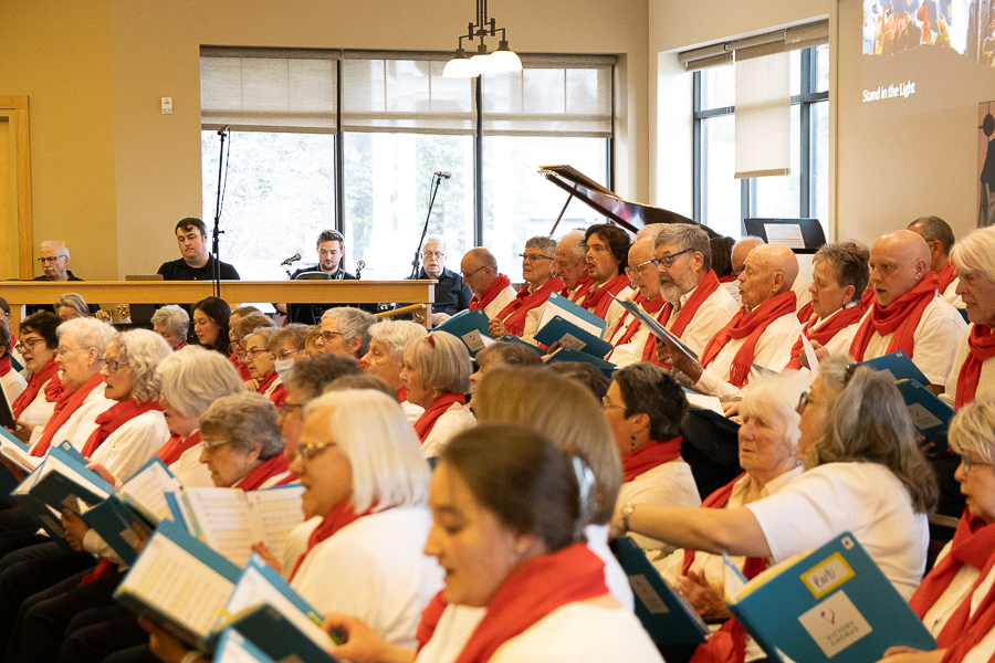 A choir rehearsal in progress with members of Victory Chorus enthusiastically singing and a conductor leading, all in a well-lit modern hall.