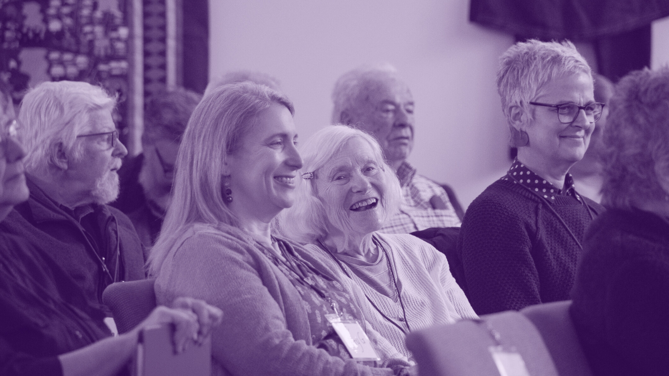 Members of Victory Chorus deeply engaged in song, with one in a wheelchair, sharing laughter and joy, in a purple-toned image that radiates warmth and inclusivity.