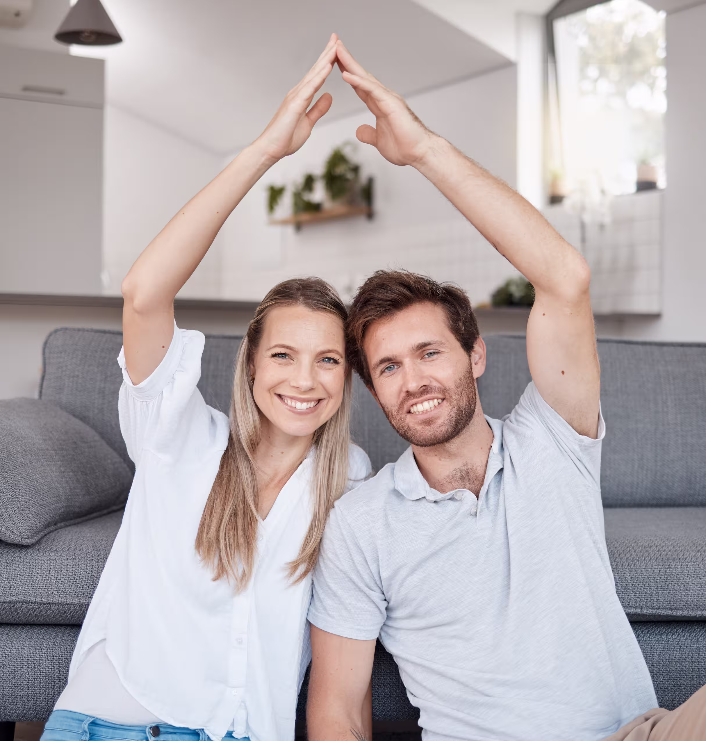 Happy couple sitting on a couch forming a house shape with their hands.