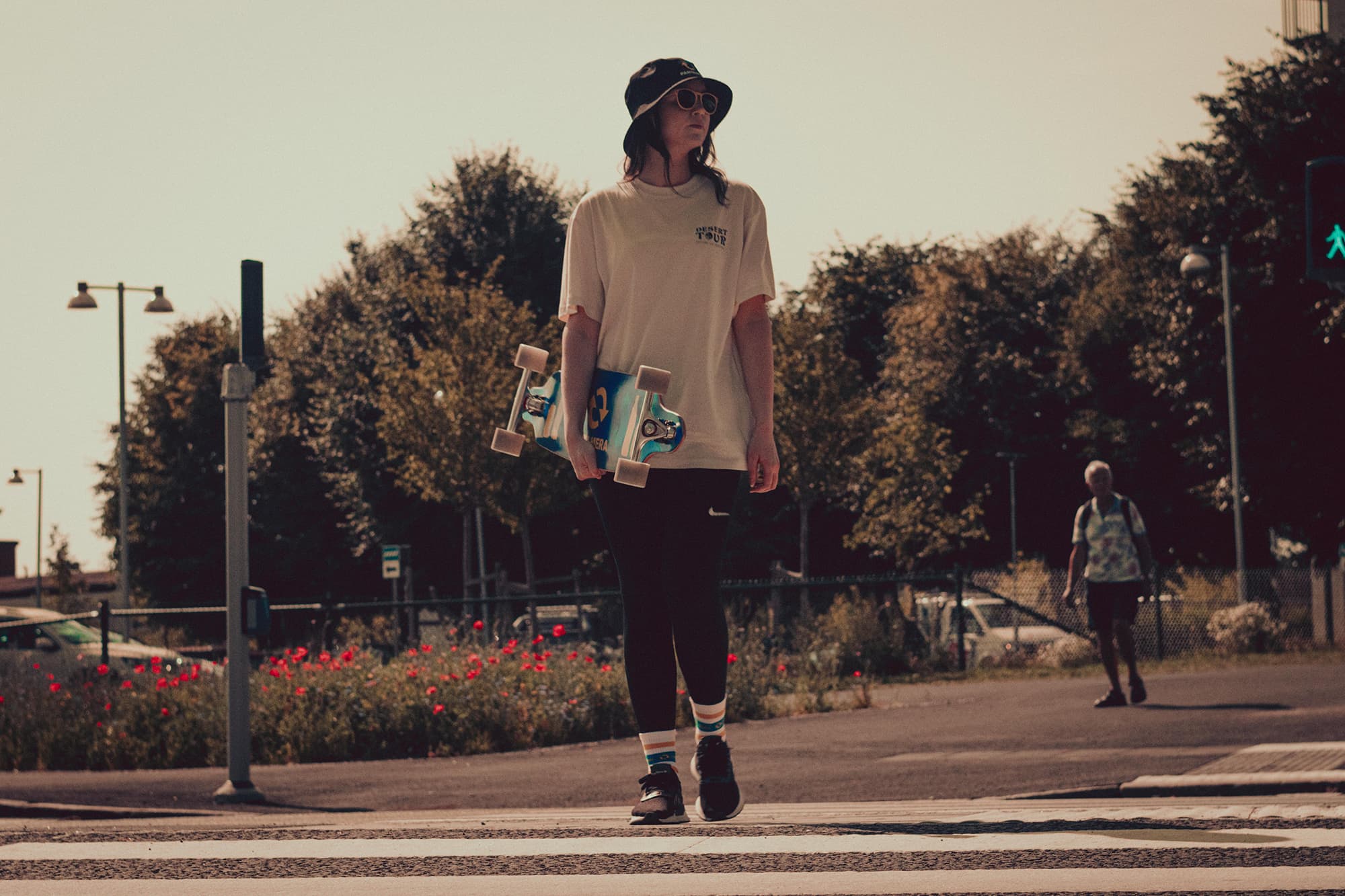Woman in sunglasses and bucket hat crossing the street carrying a longboard