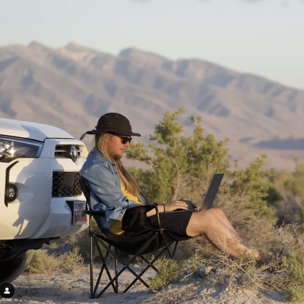 A person in a camping chair working on a laptop in the middle of the mountains