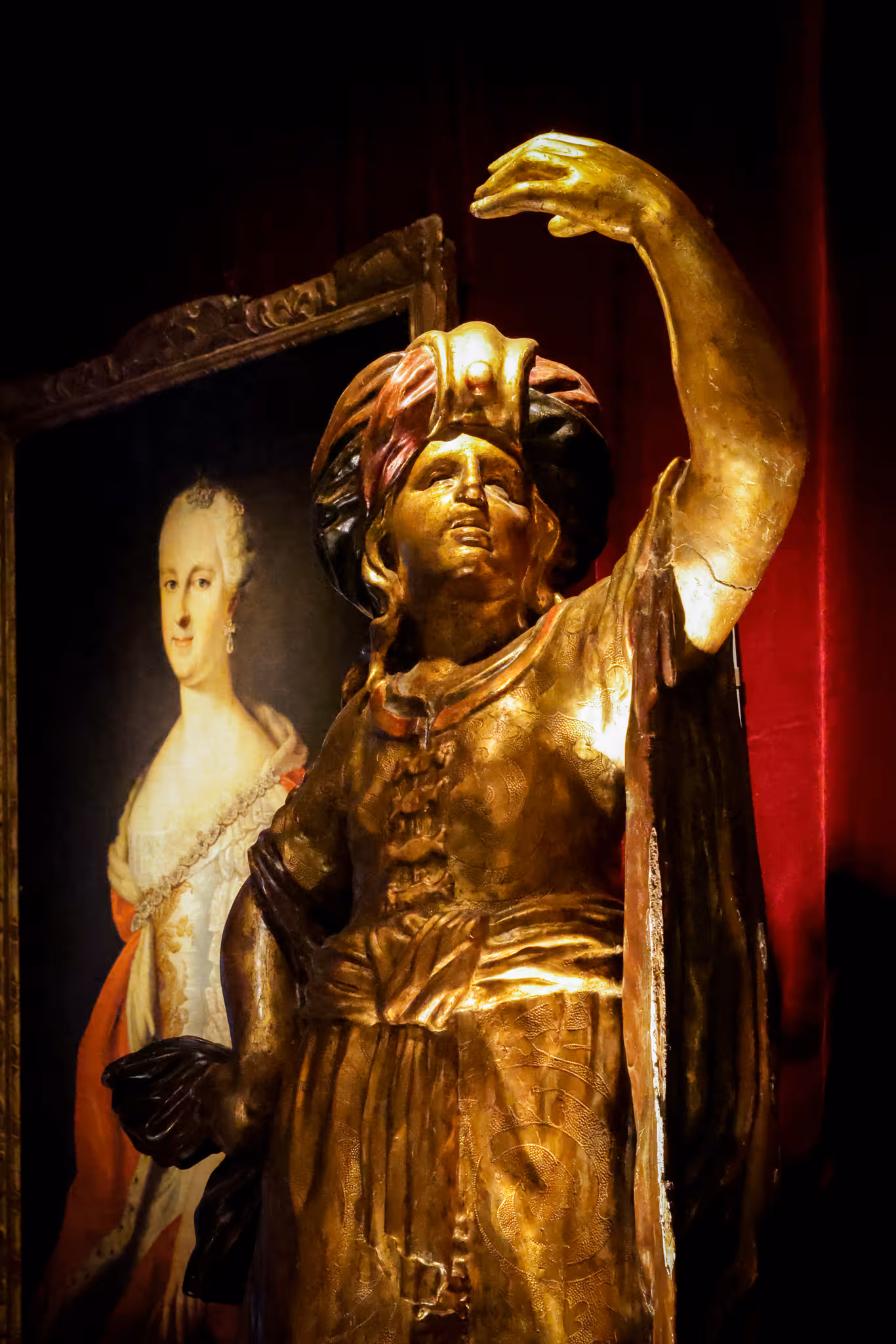 Statue dorée d’une figure vêtue d’un turbant levant le bras, devant un portrait encadré d’une femme historique en robe blanche et orange.