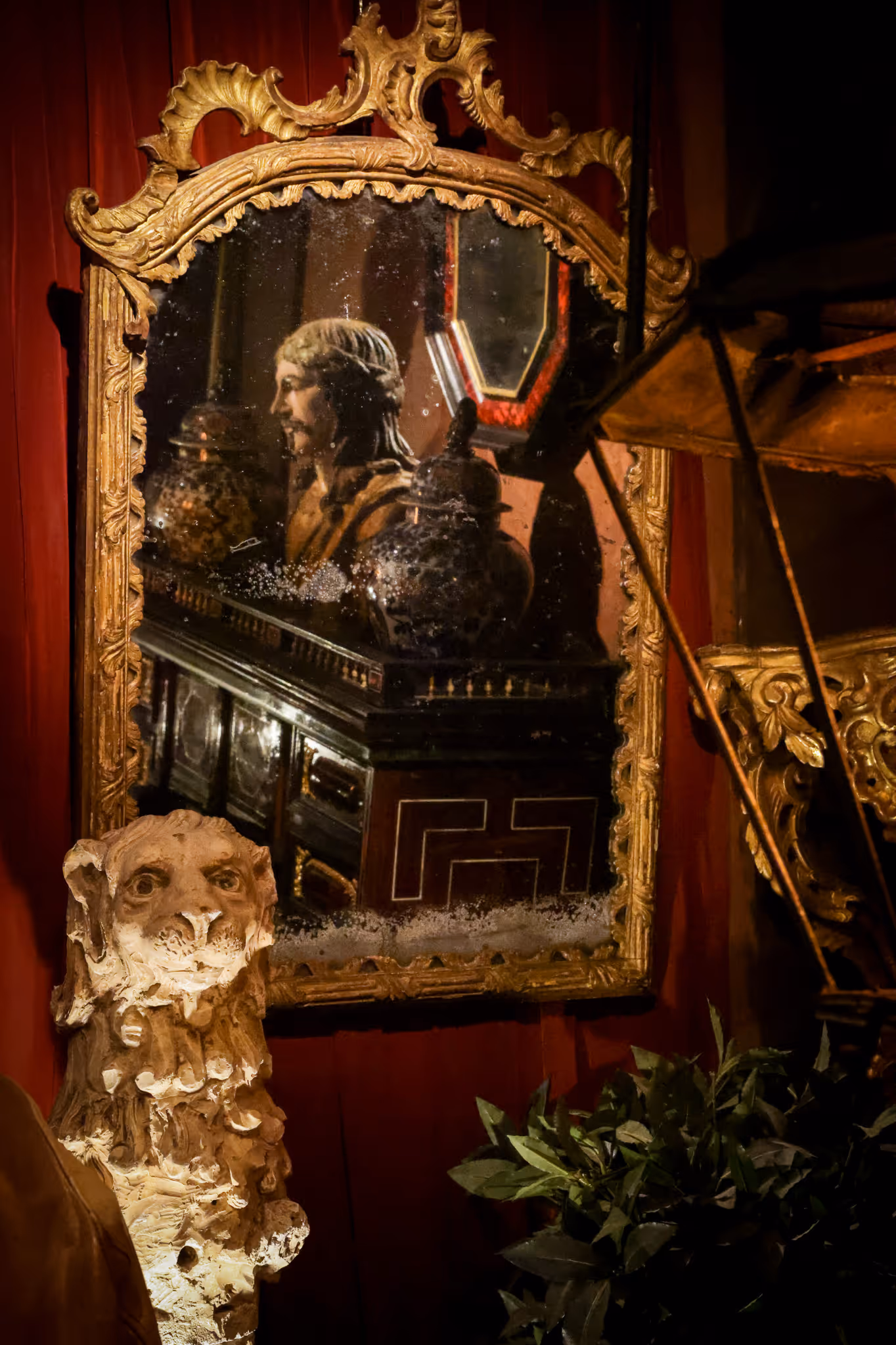 Miroir ancien encadré de bois doré reflétant une statue d’homme barbu, avec une sculpture de lion en pierre éclairée au premier plan.