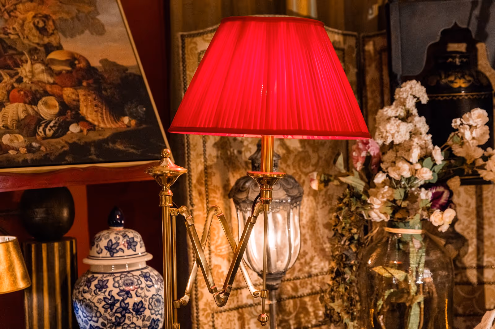 Lampe dorée avec abat-jour rouge plissé dans une pièce décorée d'un vase en porcelaine, bouquet de fleurs blanches et tableau.