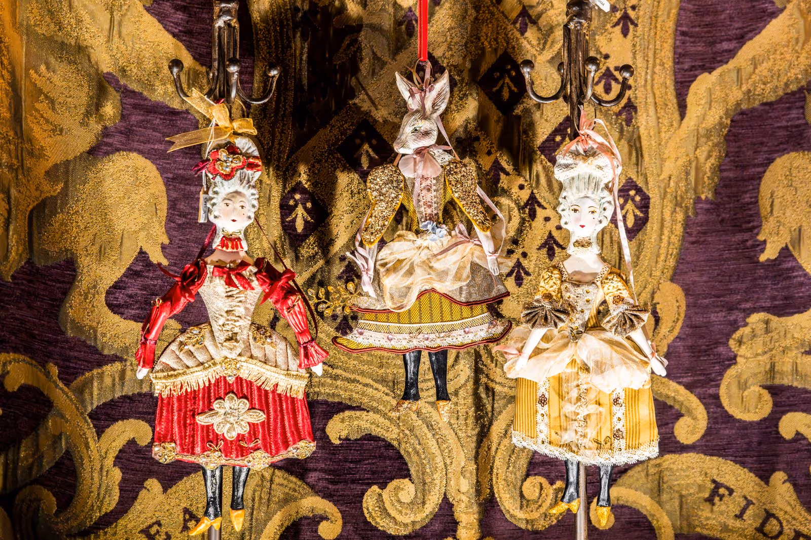 Trois figurines décoratives suspendues sur fond de tissu baroque doré et violet, deux femmes en robes d'époque et une figure mi-humaine mi-animale en tenue classique.