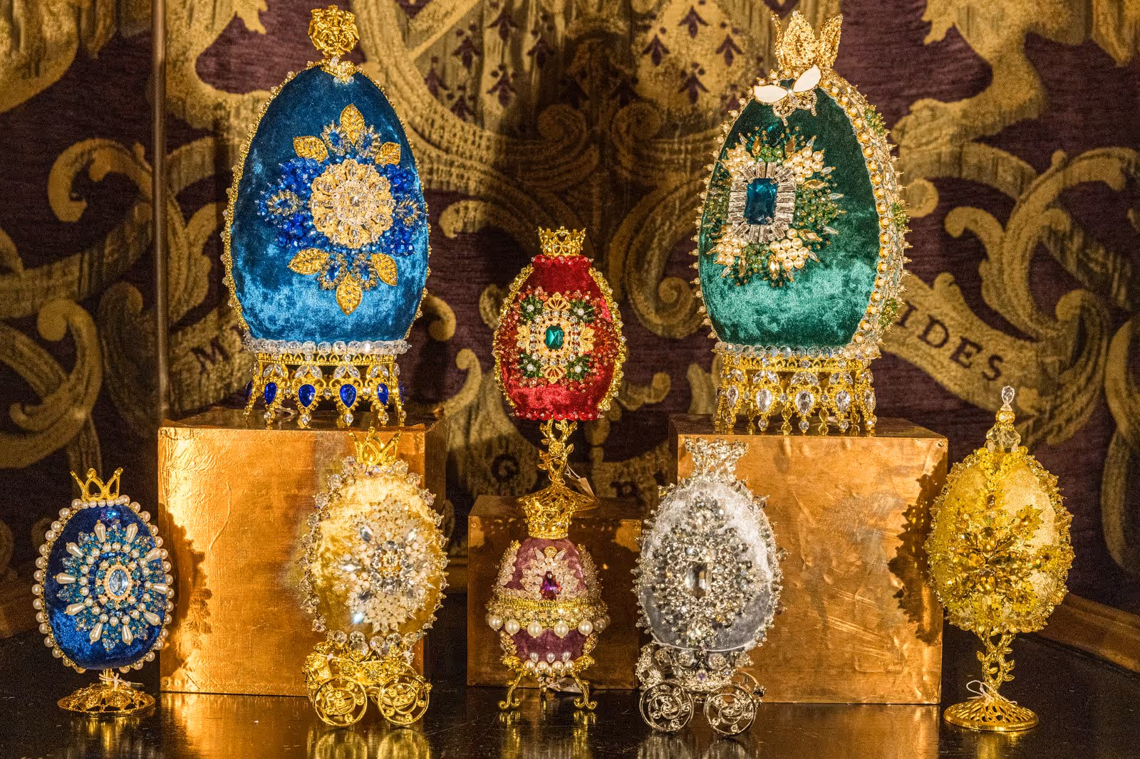 Sept œufs décoratifs de style Fabergé avec velours coloré, pierres précieuses et ornements dorés exposés sur des socles dorés.