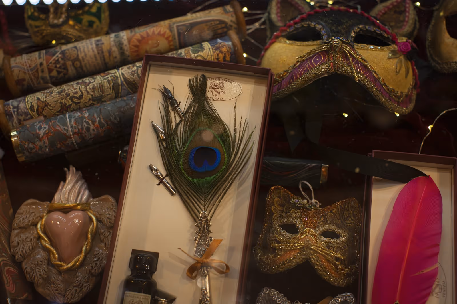 Plume de paon dans une boîte avec plumes de calligraphie, flacon d'encre, et masques vénitiens décoratifs dorés et colorés.