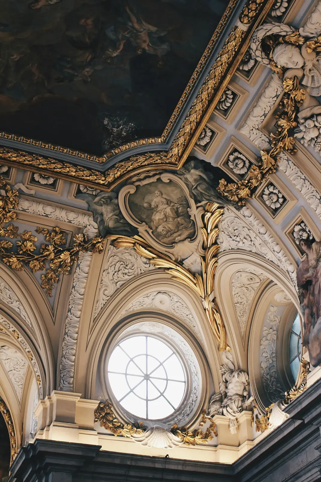 Plafond baroque avec ornement doré, fresque artistique et une fenêtre ronde laissant passer la lumière.
