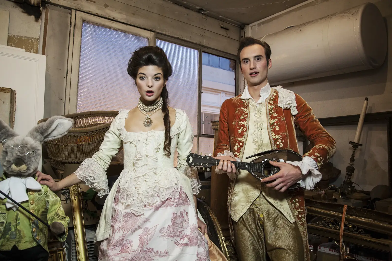 Couple en costumes d'époque dans un atelier, la femme en robe blanche brodée et le homme en veste rouge tenant un mandoline.